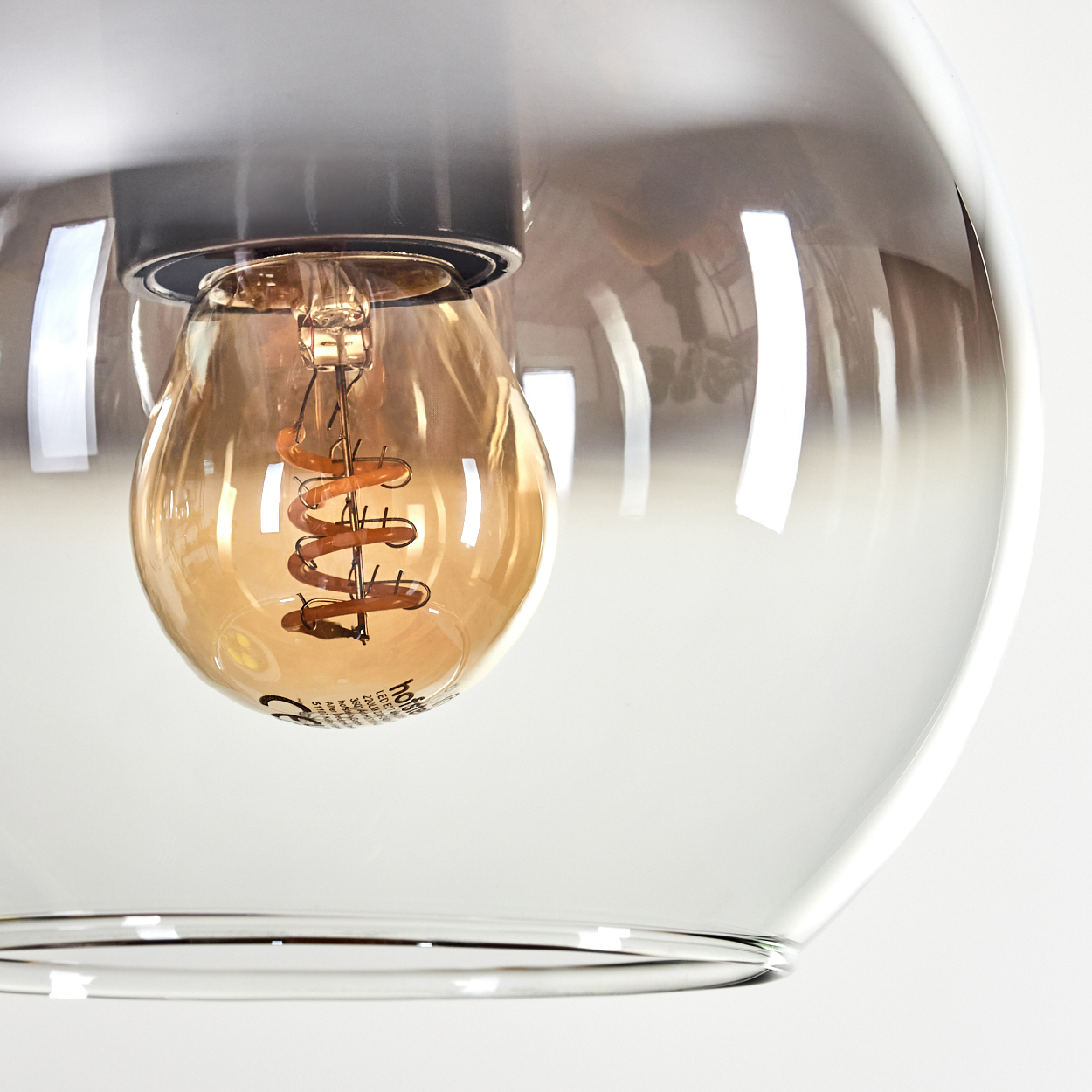 Nahaufnahme einer Leuchte mit klarem Glasschirm. Im Inneren hängt eine warme Wendelglühbirne. Der Hintergrund ist weiß.