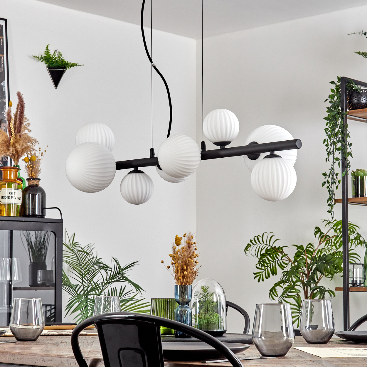 Esszimmer mit Kronleuchter, Tisch, Gläsern, Pflanzen und einem schwarzen Stuhl. Weiße Wand und schwarzes Regal im Hintergrund.