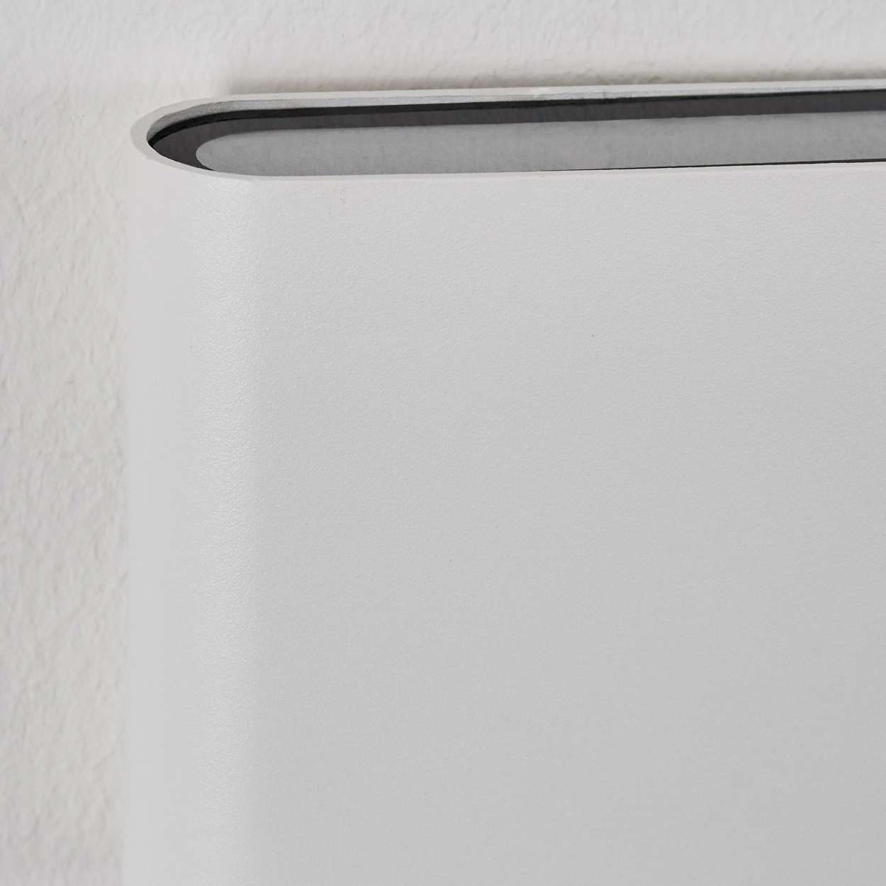 Nahaufnahme eines weißen, gebogenen Objekts vor einer weißen Wand. Das Objekt hat eine dunkelgraue Oberseite und eine glatte, helle Oberfläche.