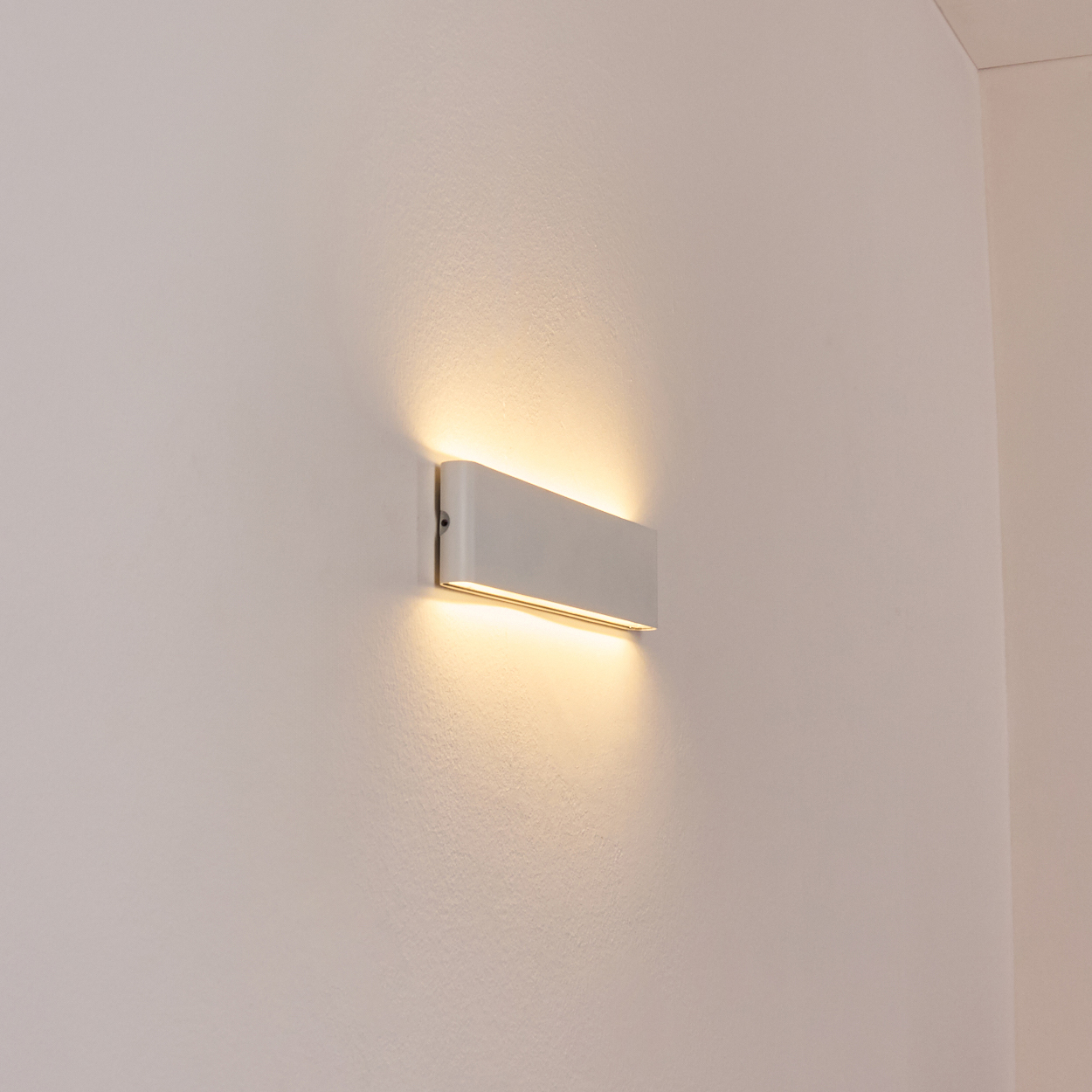 Eine weiße, rechteckige Wandleuchte ist an einer weißen Wand montiert. Die Lampe leuchtet und wirft einen warmen Schein.