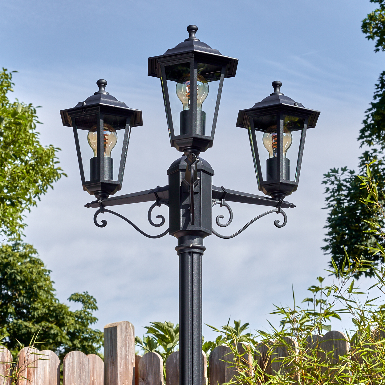 Eine schwarze Laterne mit drei Lampen, jede mit einer Glühbirne. Sie steht vor blauem Himmel, Holzzaun und grünen Pflanzen.