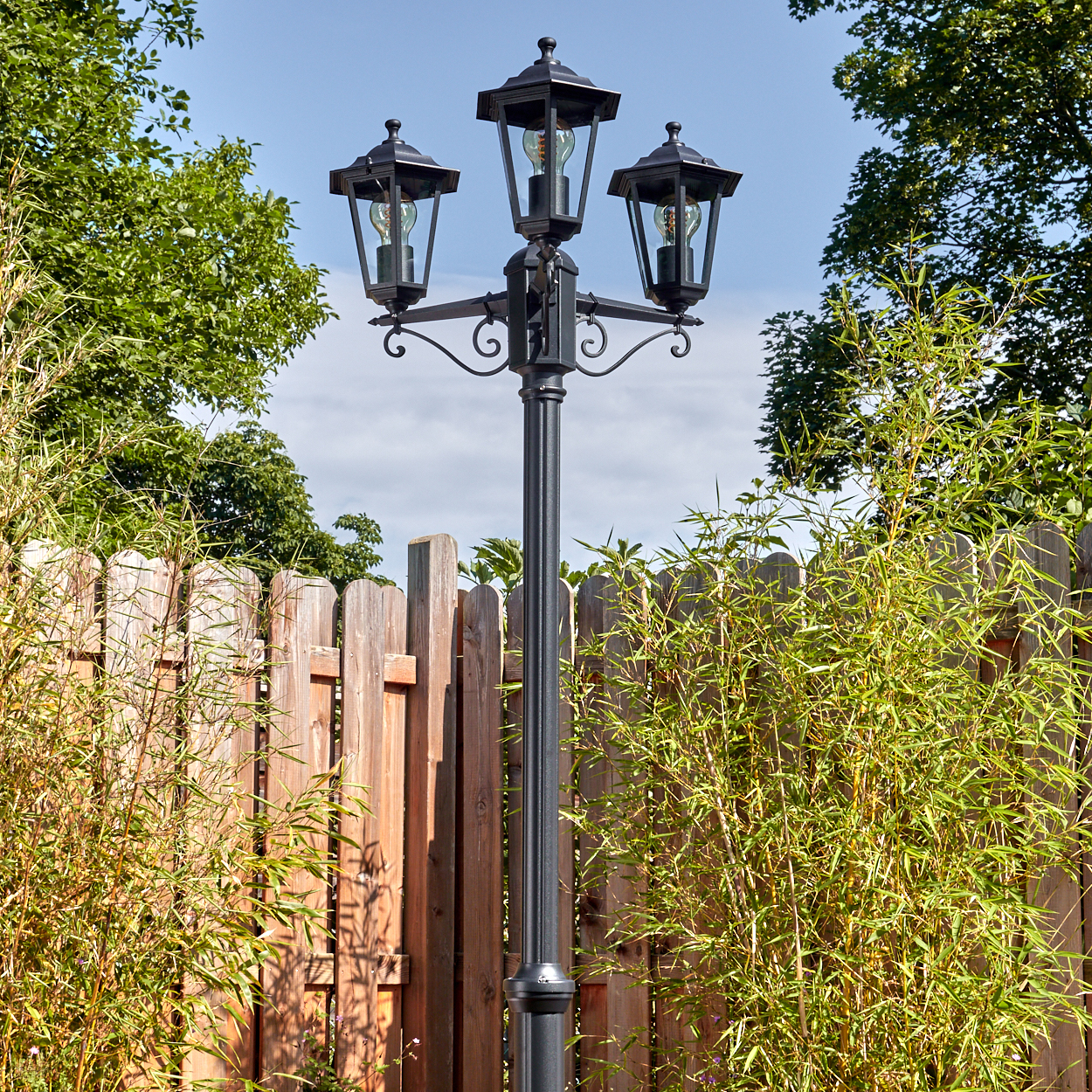Eine schwarze Laterne mit drei Lampen steht vor einem Holzzaun, umgeben von grünem Bambus und Bäumen, und blauem Himmel.