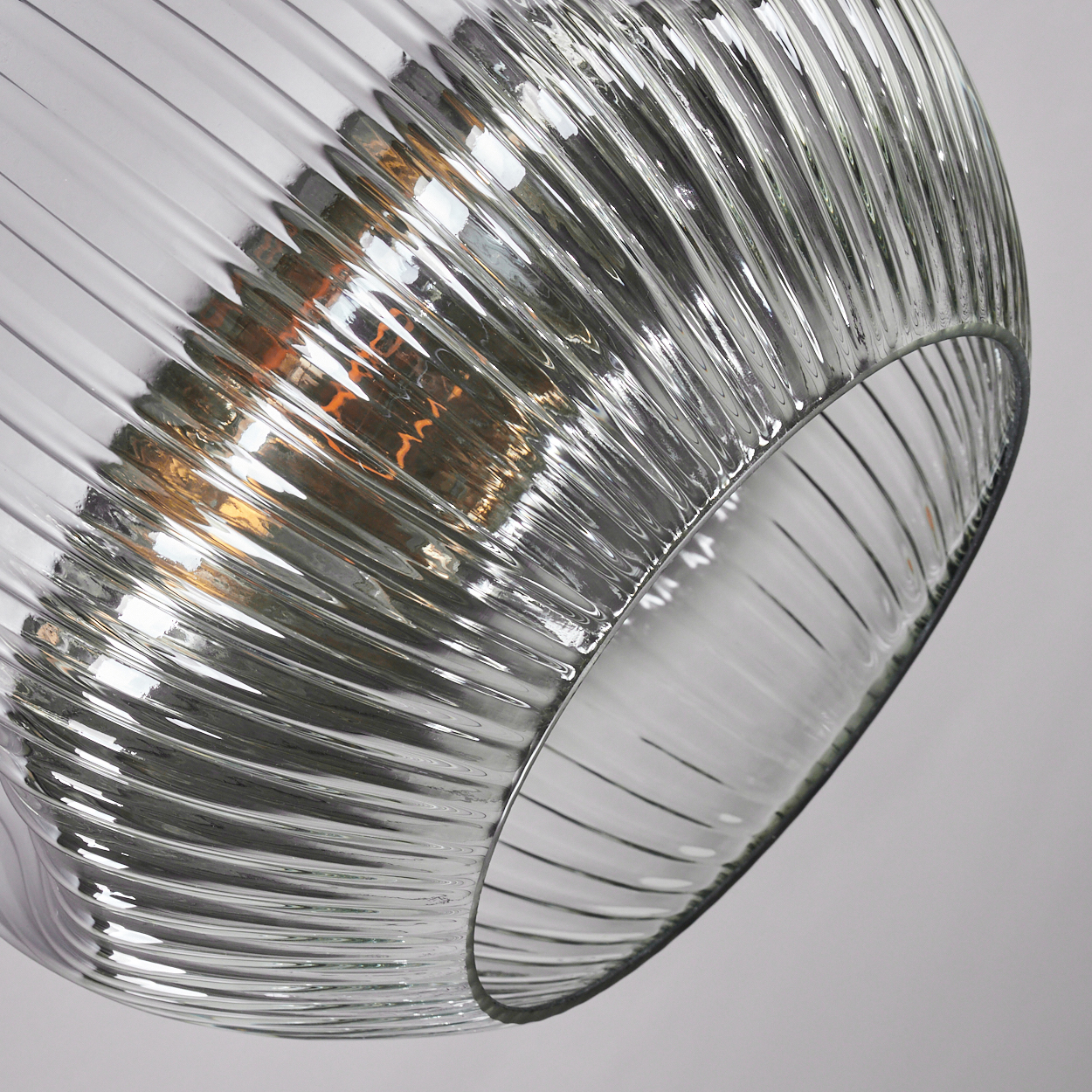 Nahaufnahme eines klaren, gerippten Glaslampenschirms. Licht reflektiert sich im Glas, mit einem warmen Schein im Inneren. Der Hintergrund ist weiß.