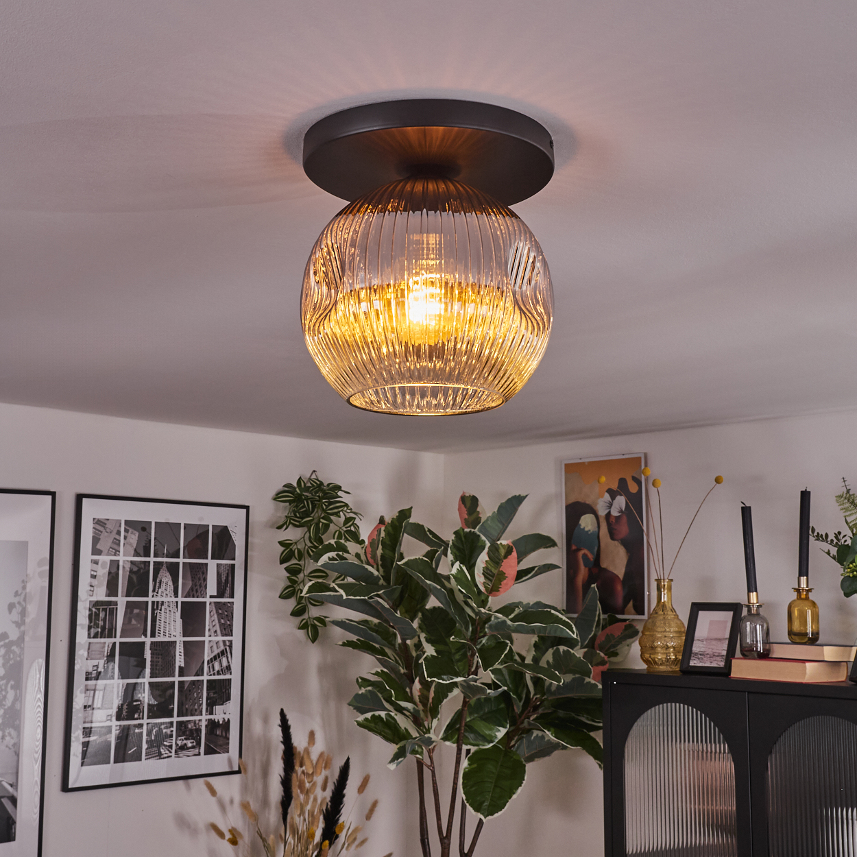 Eine dekorative Deckenlampe mit Glasschirm leuchtet in einem Raum. Pflanzen und Kunstwerke sind im Hintergrund zu sehen.