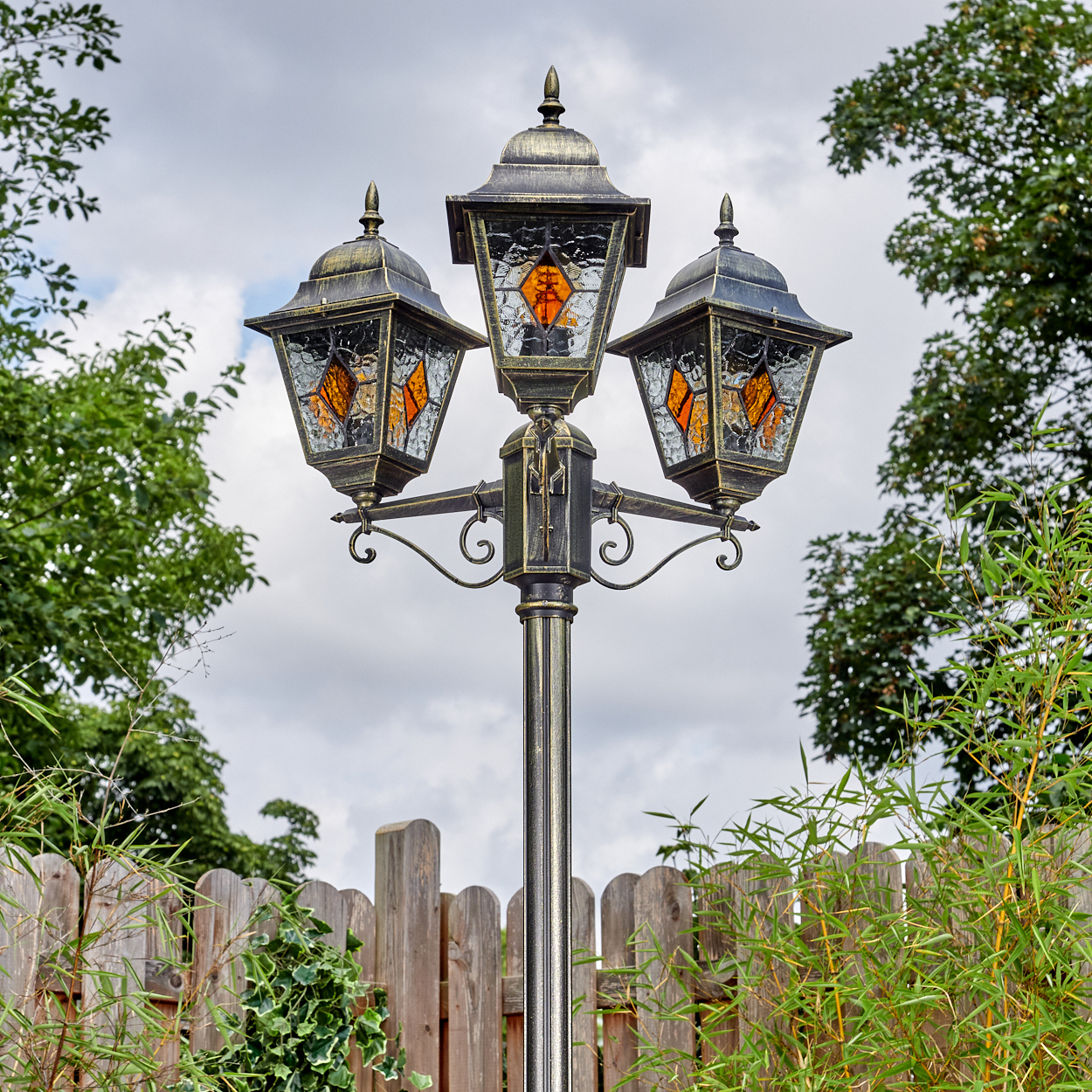 Eine verzierte Laternenpfahl mit drei Lampen vor bewölktem Himmel. Ein Holzzaun und grünes Laub sind im Vordergrund und an den Seiten.