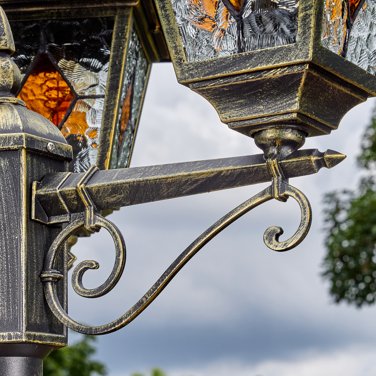 Nahaufnahme einer dekorativen Laterne. Sie hat Buntglasscheiben, filigrane Metallarbeiten mit Schwarz-Gold-Finish und einen bewölkten Himmel im Hintergrund.