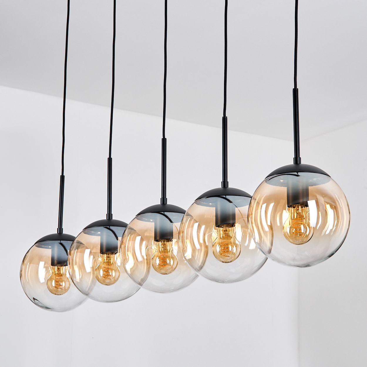 Fünf hängende Kugelleuchten mit bernsteinfarbenen Glühbirnen. Schwarze Armaturen. Weiße Decke und Wand. Klare Glasschirme mit schwarzen Oberteilen.