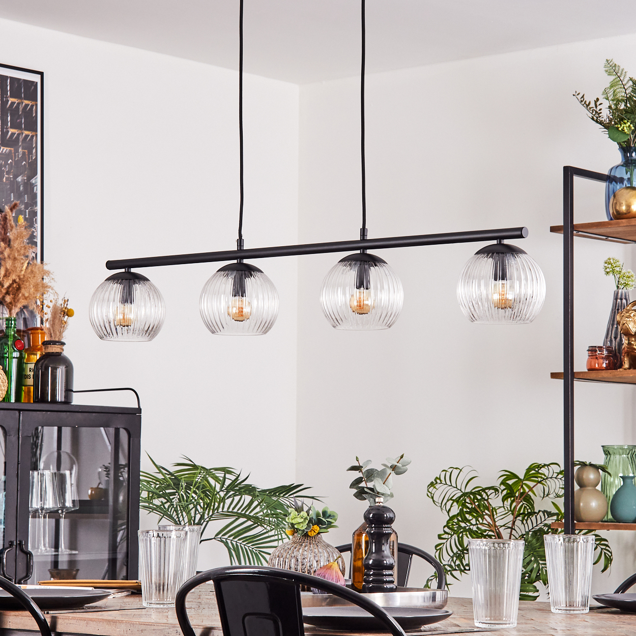 Ein Esszimmer mit Holztisch, schwarzen Stühlen und einer schwarzen Leuchte mit vier Glaskugeln. Pflanzen und Dekorationen sind vorhanden.