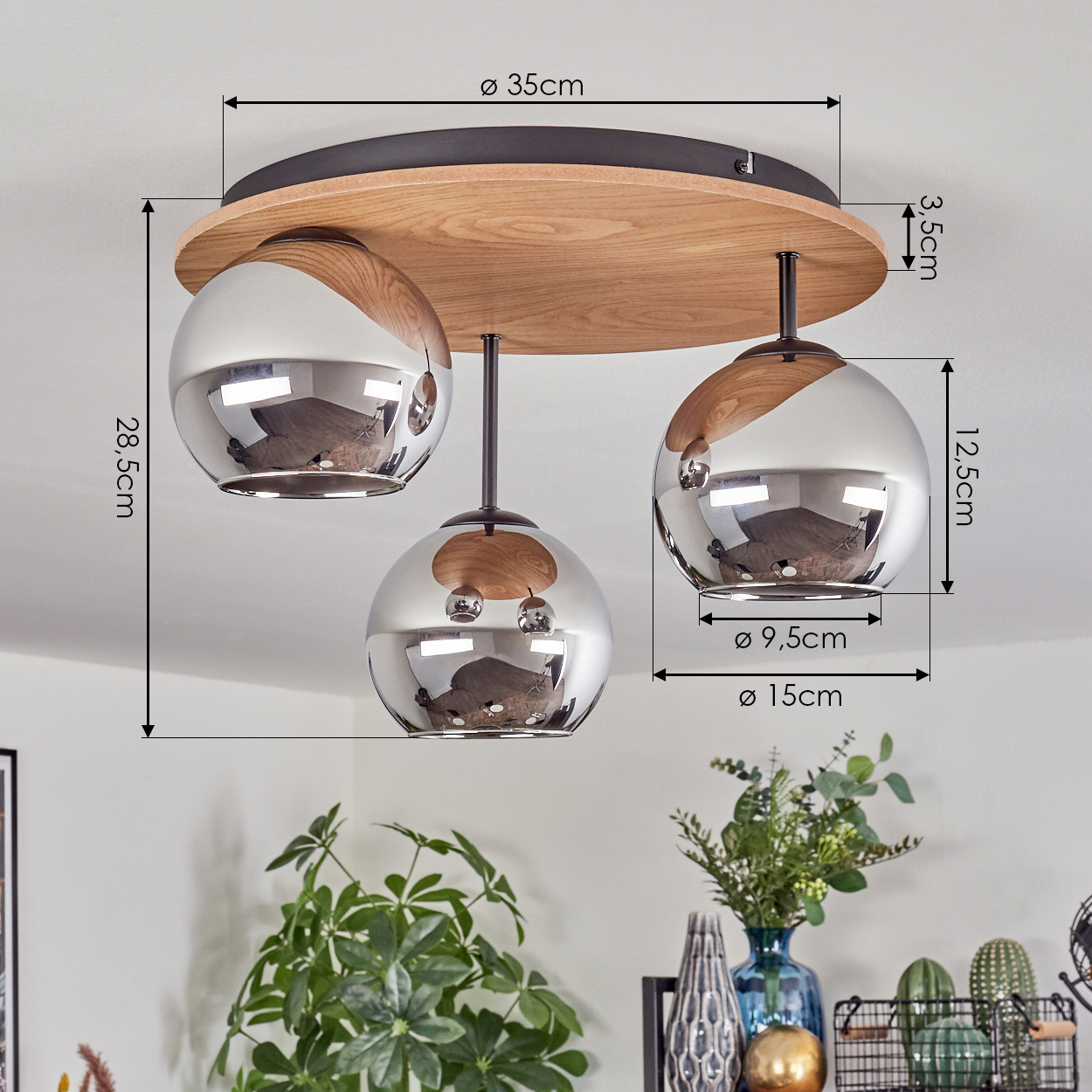 Eine moderne Deckenlampe mit drei kugelförmigen Schirmen. Die Lampe hat einen Holzsockel und silberne Schirme, mit Pflanzen im Hintergrund.