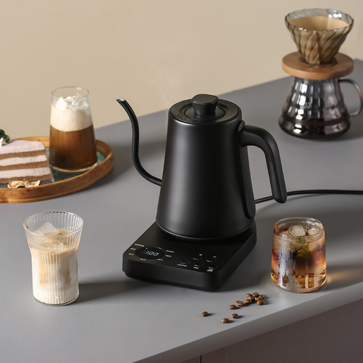 Ein schwarzer Wasserkocher mit Kaffee und Getränken auf einer grauen Oberfläche. Im Hintergrund sind Kuchen und eine Kaffeemaschine.