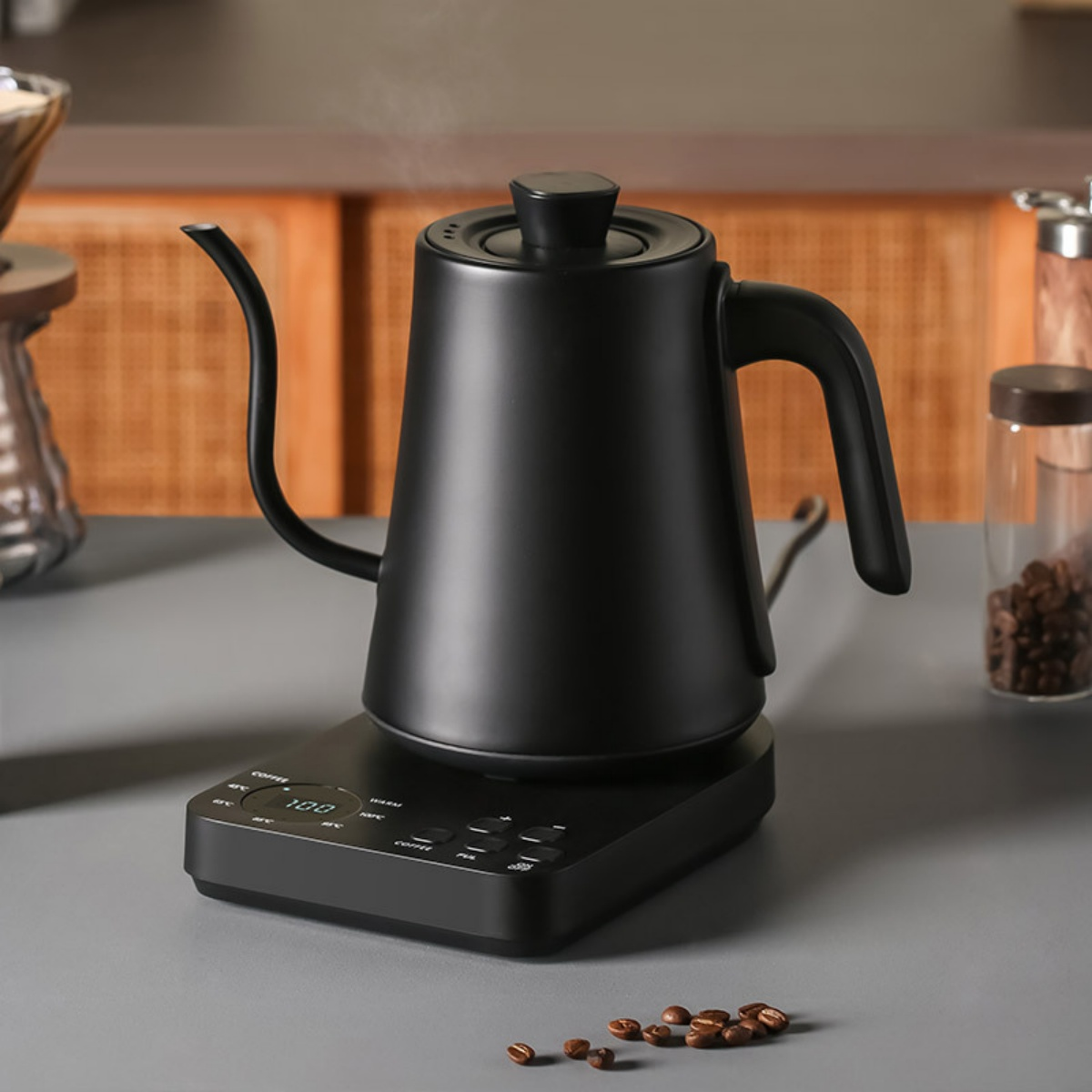 Ein schwarzer Wasserkocher mit aufsteigendem Dampf steht auf einem schwarzen Sockel. Kaffeebohnen und ein Glas stehen im Hintergrund.