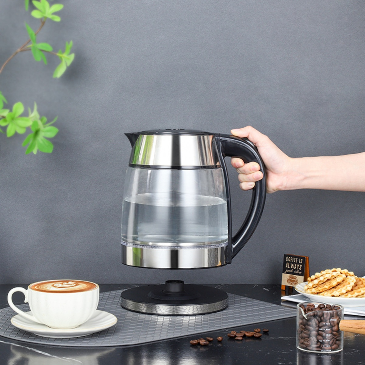 Eine Hand hält einen Glaskessel mit Wasser. Auf dem Tisch stehen eine Tasse Kaffee, Waffeln und Kaffeebohnen.