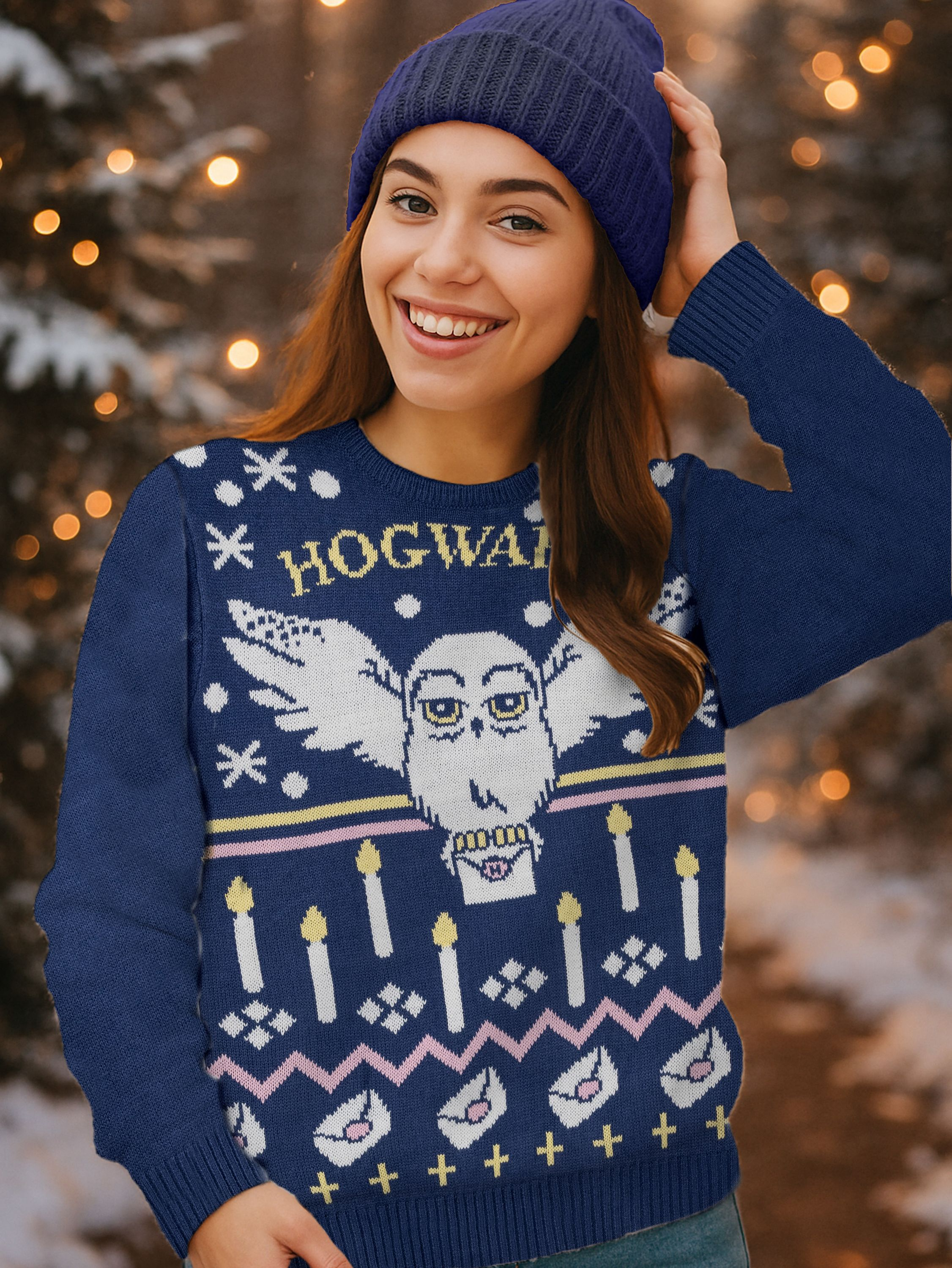Lächelnde Frau in blauem Beanie und Pullover mit Hogwarts-Design, draußen mit Bokeh-Lichtern im Hintergrund.