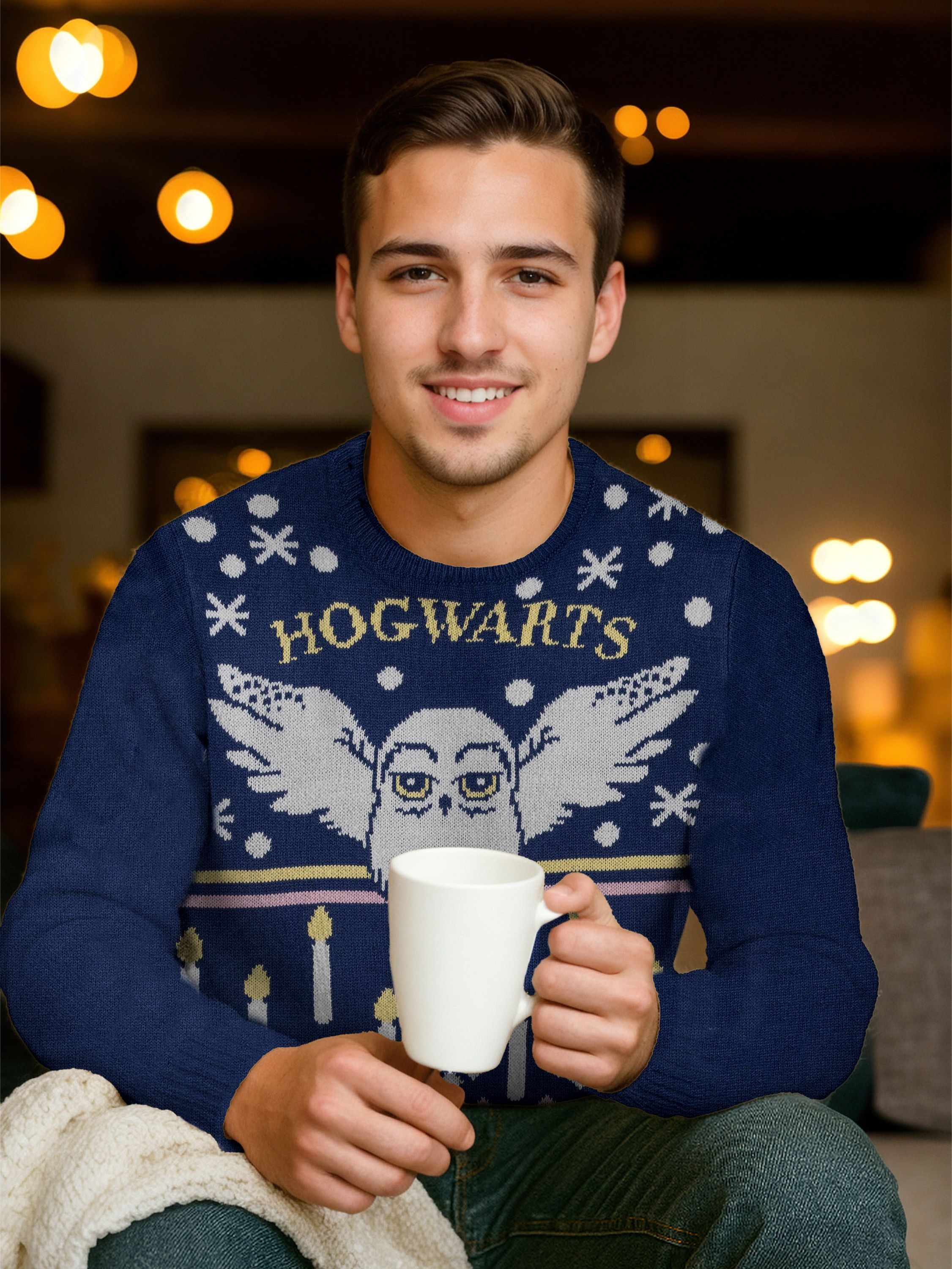 Ein Mann lächelt und hält eine weiße Tasse. Er trägt einen blauen Hogwarts-Pullover. Hintergrund mit verschwommenen Lichtern.