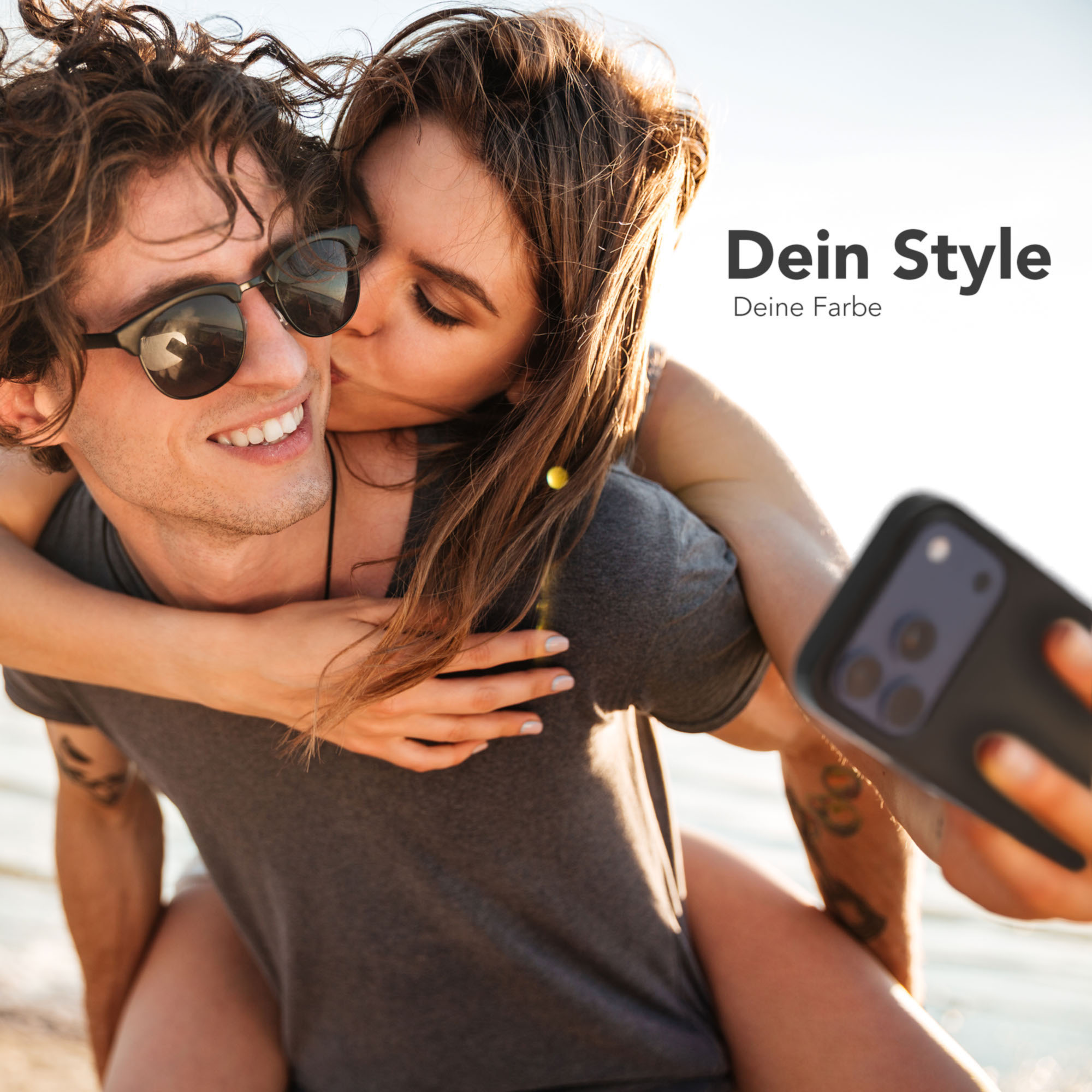 Ein Mann mit Sonnenbrille lächelt, während eine Frau ihn auf die Wange küsst. Sie machen ein Selfie.