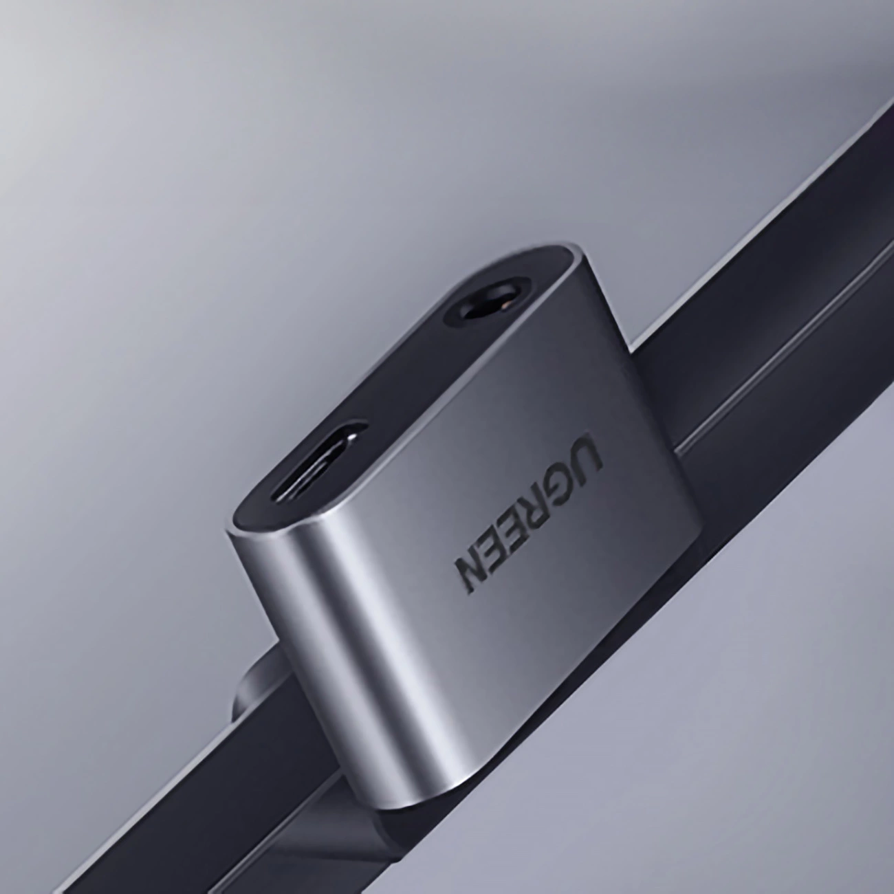 Pokazano srebrny adapter UGREEN. Posiada port USB-C, gniazdo słuchawkowe i jest zamontowany na ciemnej powierzchni na jasnym tle.