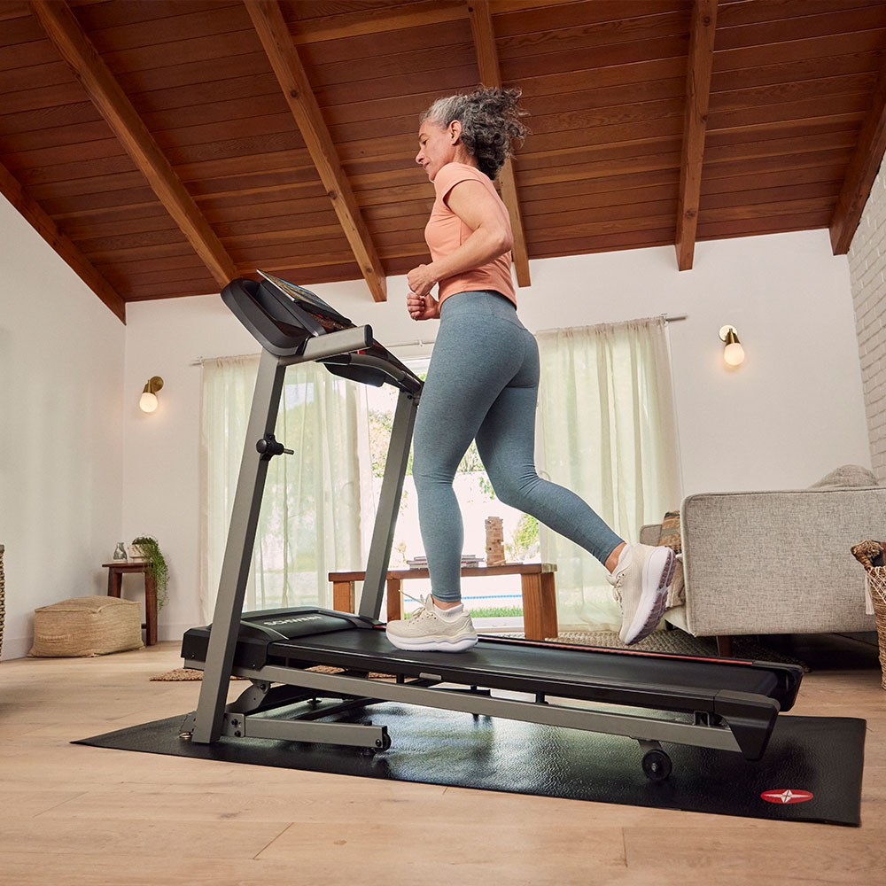 A woman runs on a treadmill. She wears leggings and a top, in a home setting. The treadmill is on a mat.