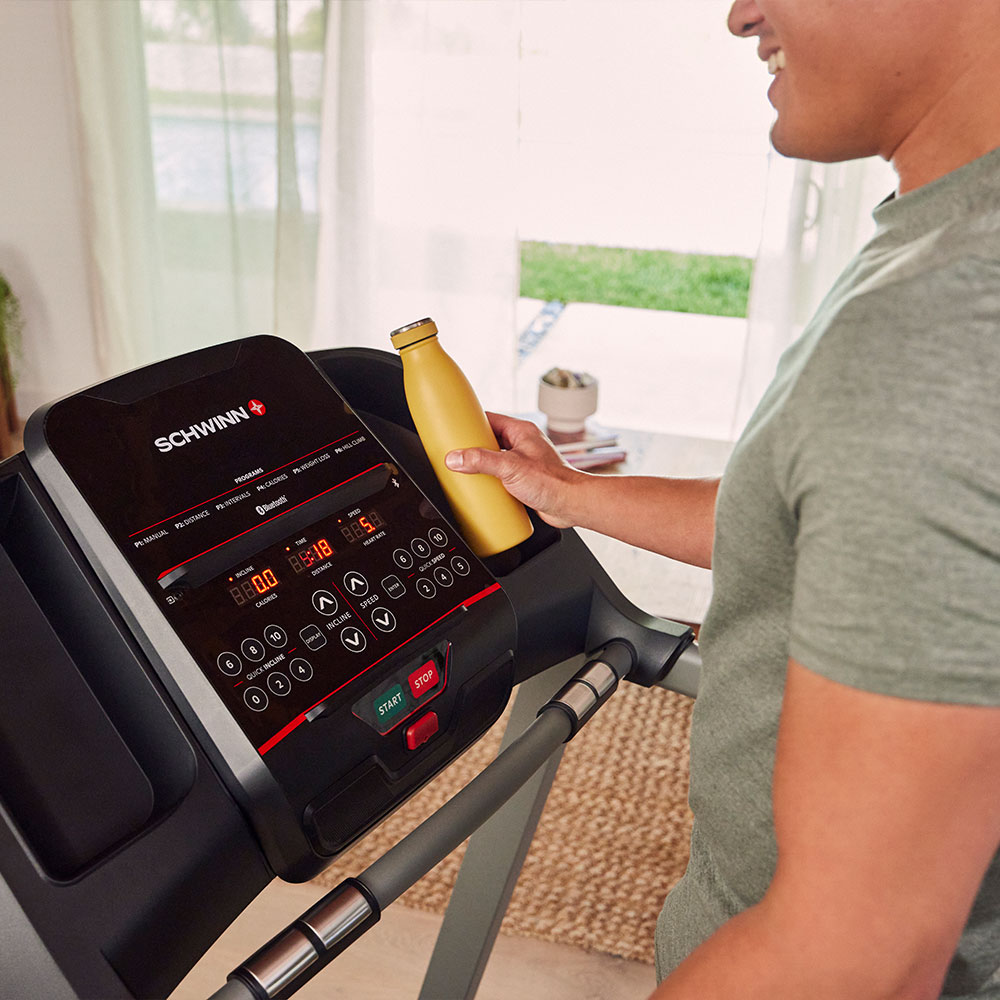 A man holds a yellow water bottle on a treadmill. The treadmill displays stats. Behind is a window and a small plant.