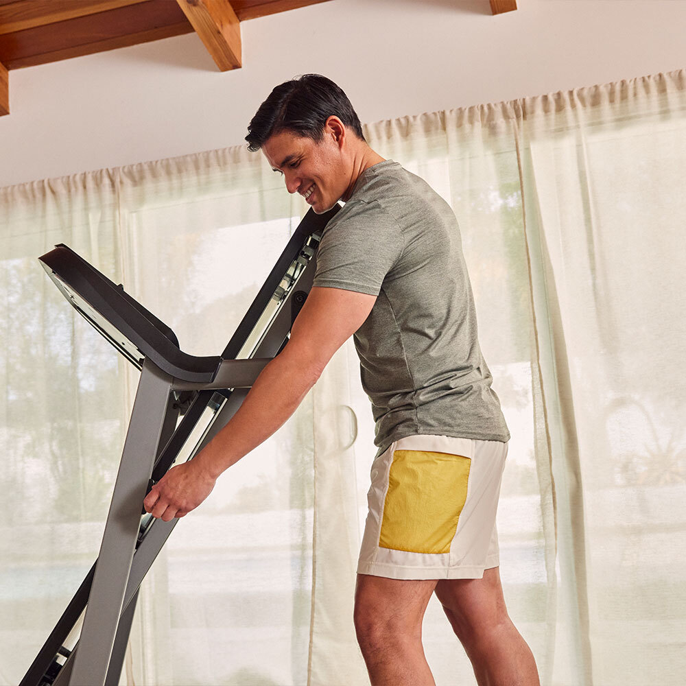 A man smiles while adjusting a treadmill. He wears athletic clothes. Background has curtains and wooden beams.