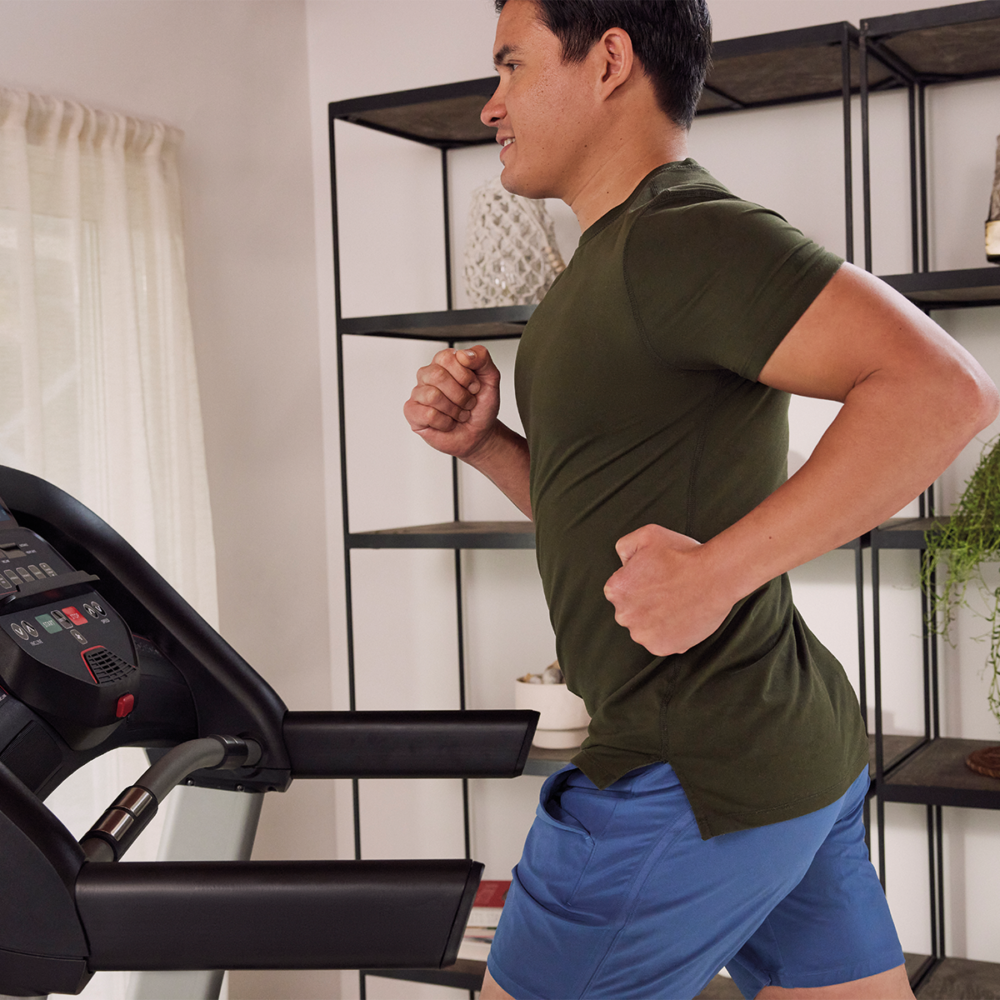 A man runs on a treadmill. He wears blue shorts and a green shirt. A shelf and curtain are in the background.