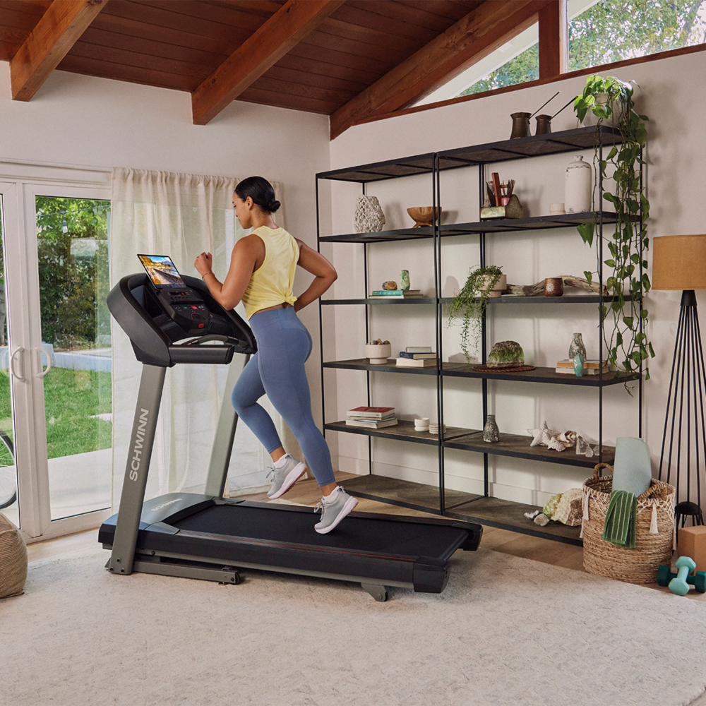 A woman runs on a treadmill. She wears blue leggings and a yellow top. The room has a shelf, a window, and a light.