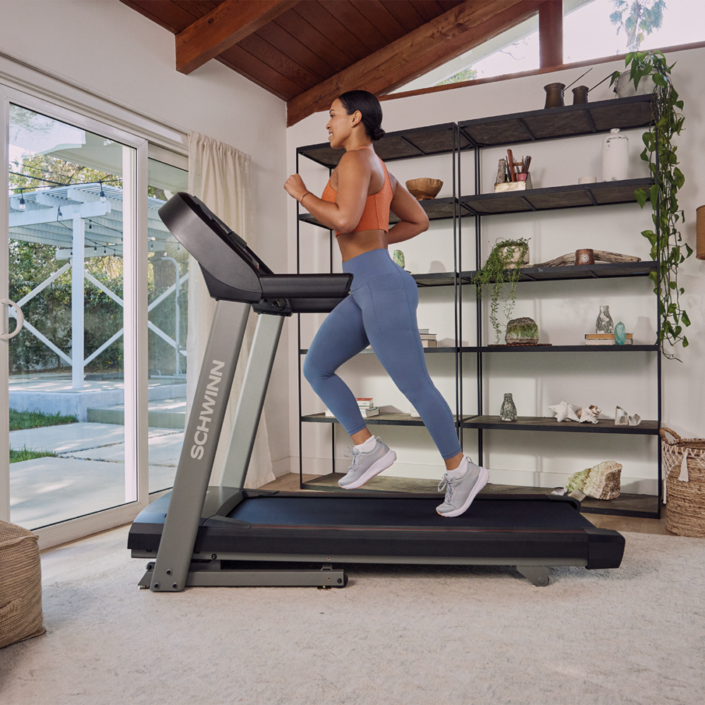 A woman runs on a treadmill. She wears activewear. The treadmill is in a bright room with shelves and a window.