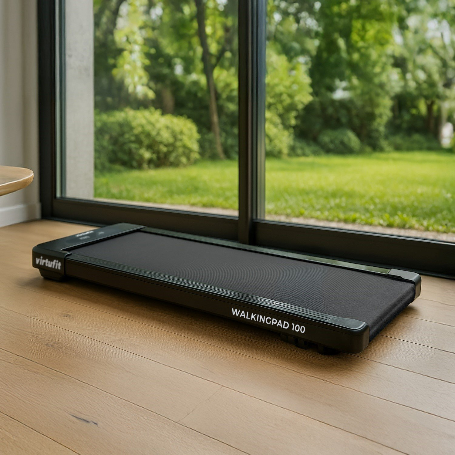 Un walkingpad nero è appoggiato su un pavimento in legno vicino a una finestra. Fuori dalla finestra c'è un prato verde e alberi.