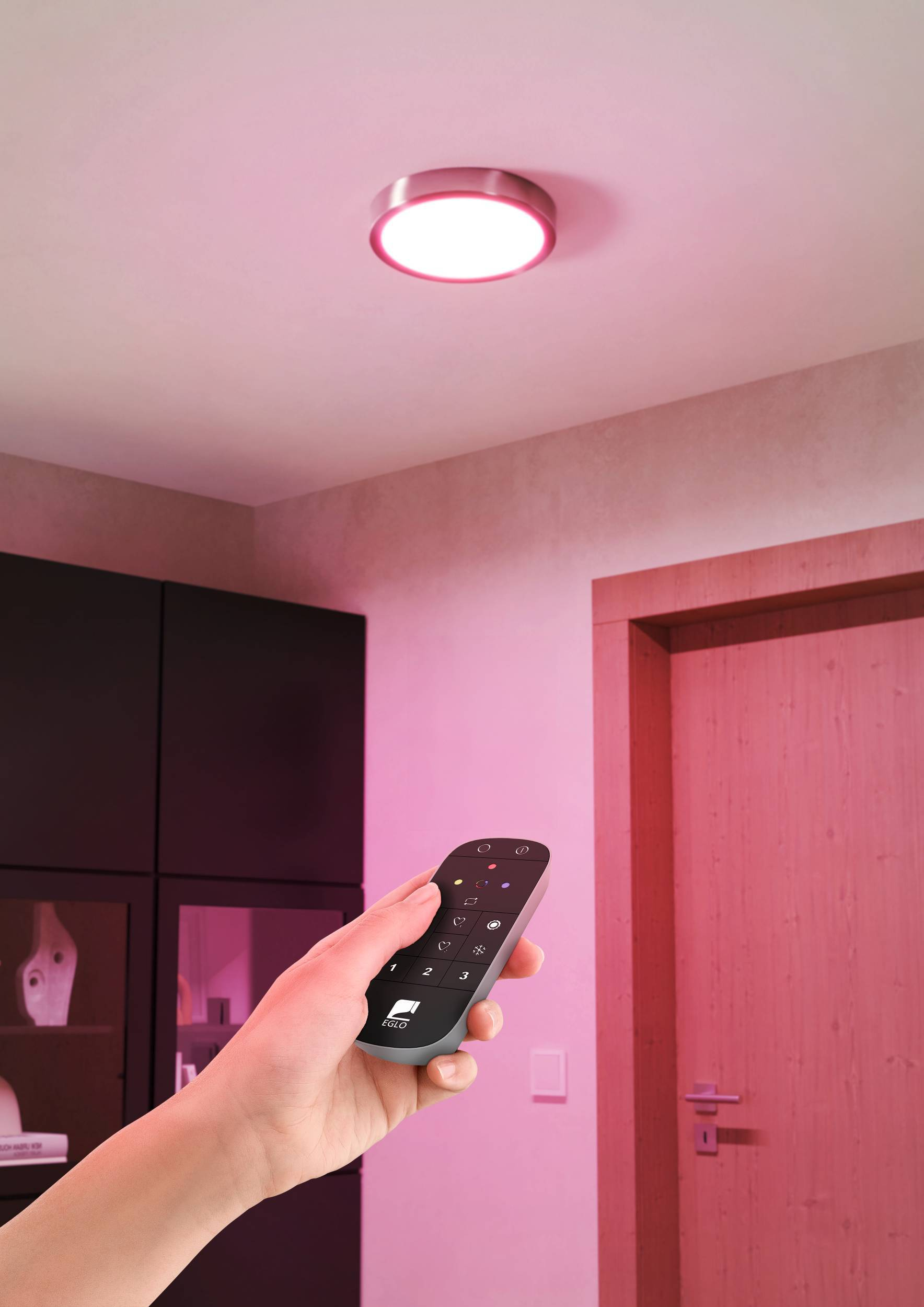 Una mano tiene un telecomando che punta a una plafoniera. La stanza è illuminata di rosa, si vede una porta.