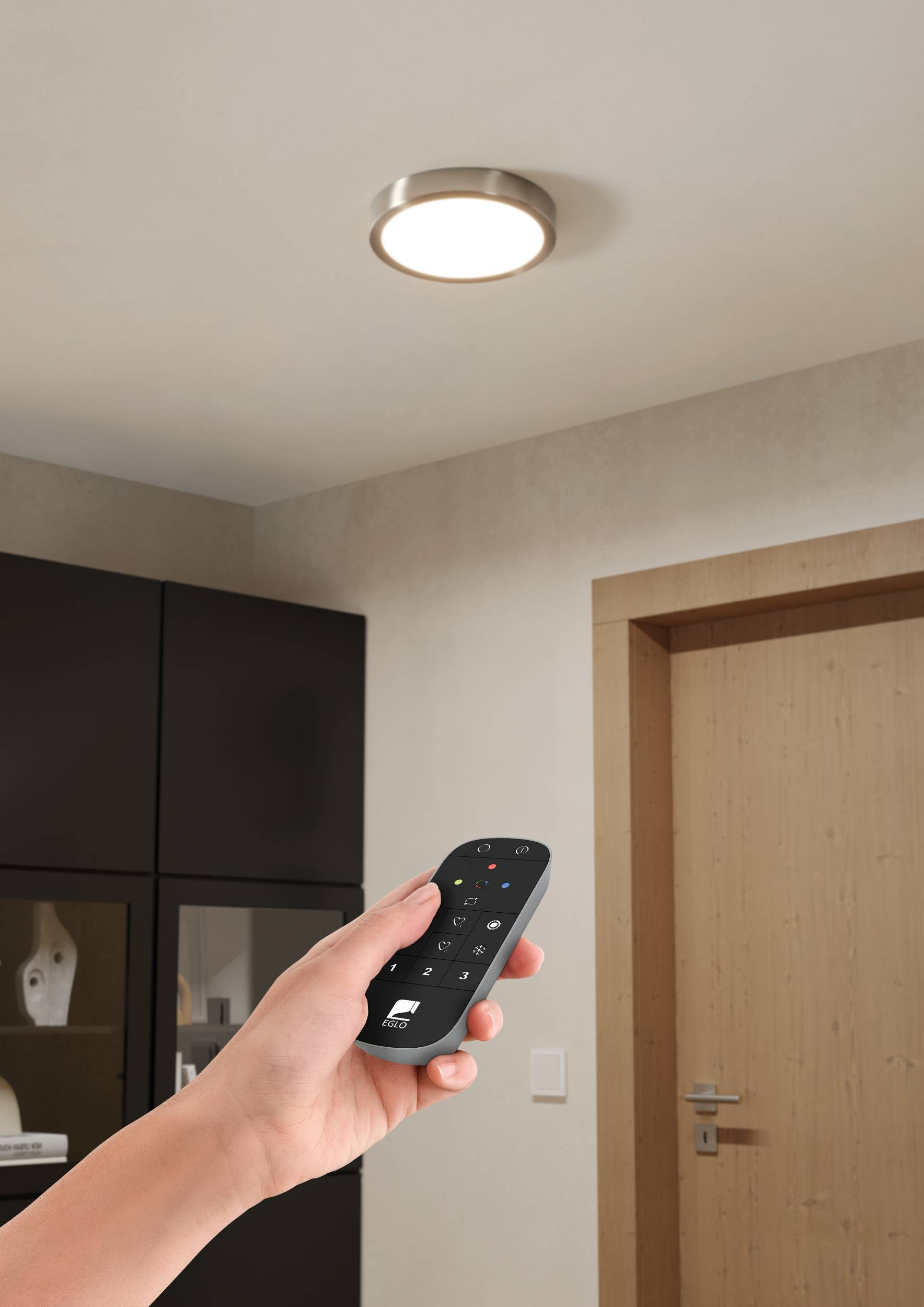 Mano che tiene un telecomando che punta alla luce del soffitto. Armadi neri e porta in legno sullo sfondo.