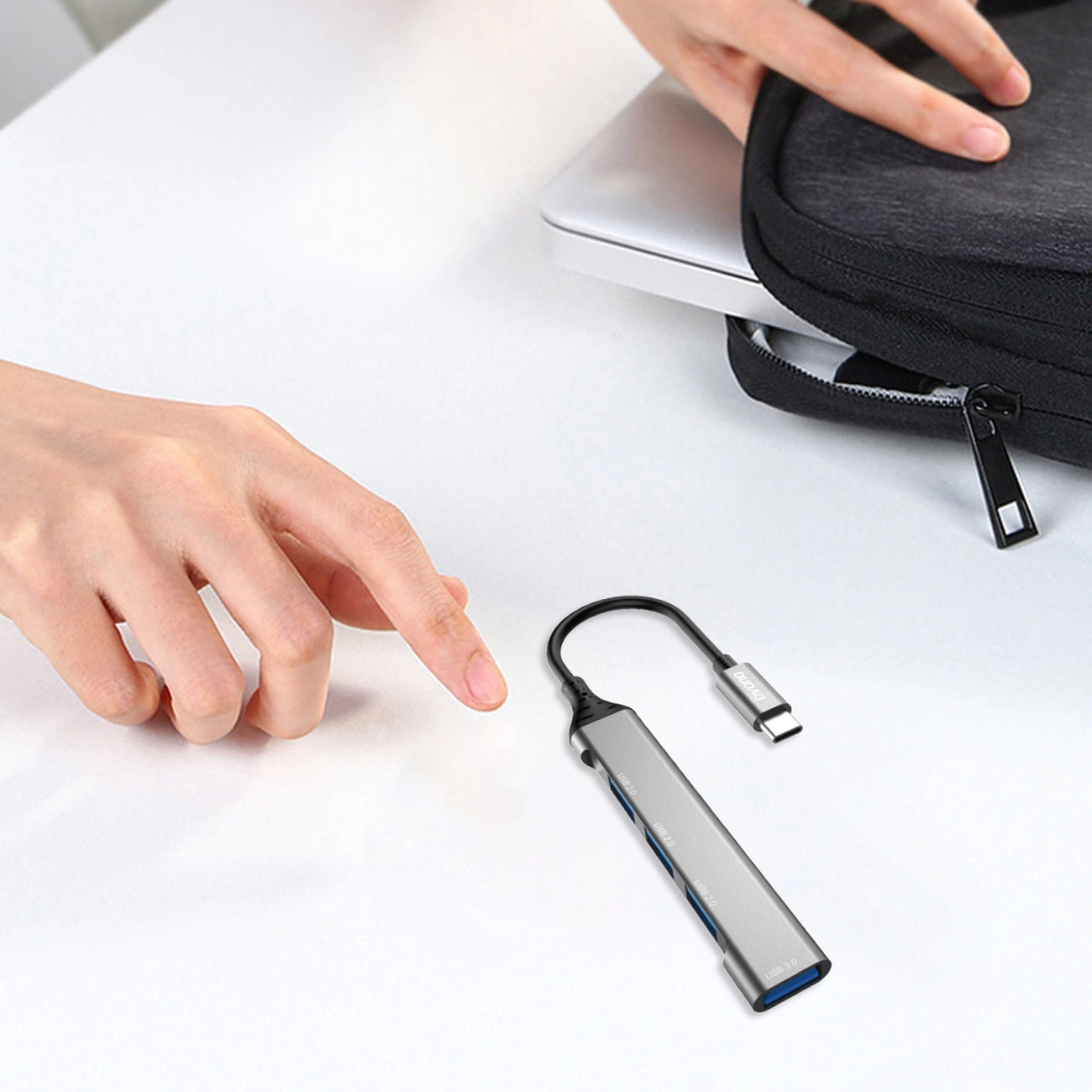 Una mano indica un hub USB argentato su una superficie bianca. Un laptop e una custodia nera sono sullo sfondo.