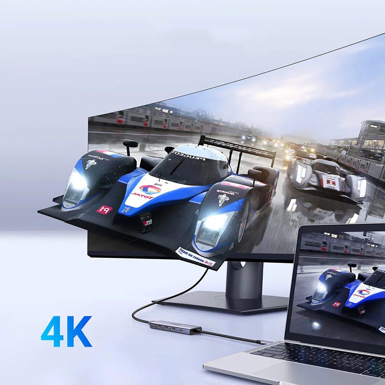 Monitor wyświetla scenę z wyścigami samochodowymi, połączony z laptopem za pomocą huba. Widoczne jest słowo 4K. Scena ma miejsce w dzień.
