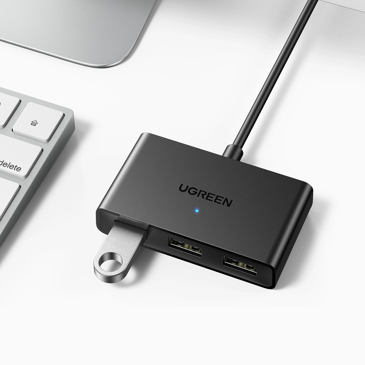 Un hub USB UGREEN nero con anello in metallo argentato, collegato a un cavo nero. Tastiera e laptop.