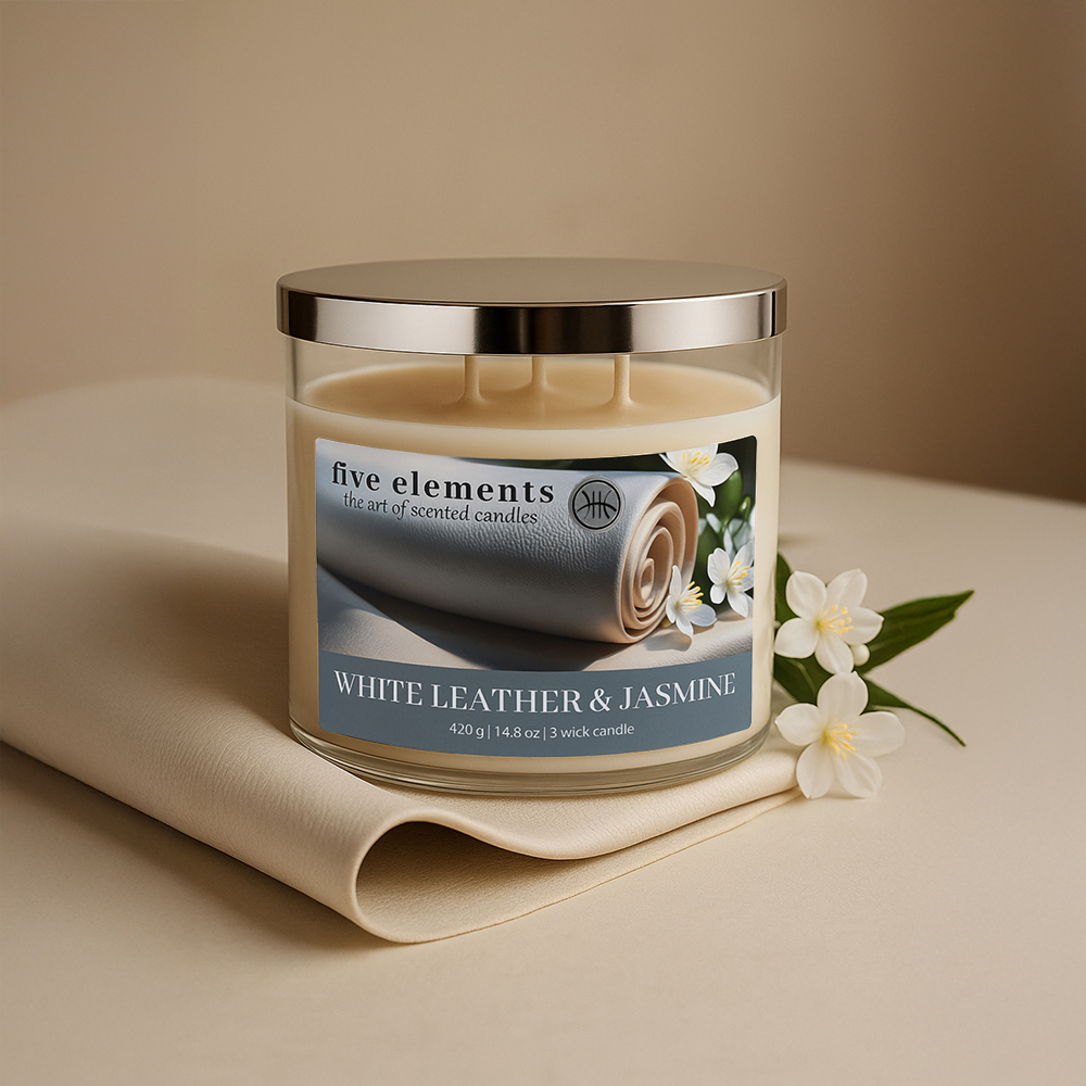 Eine Kerze mit silbernem Deckel steht auf einem beigen Tuch. Auf dem Etikett steht 'White Leather & Jasmine'. Blumen liegen daneben.