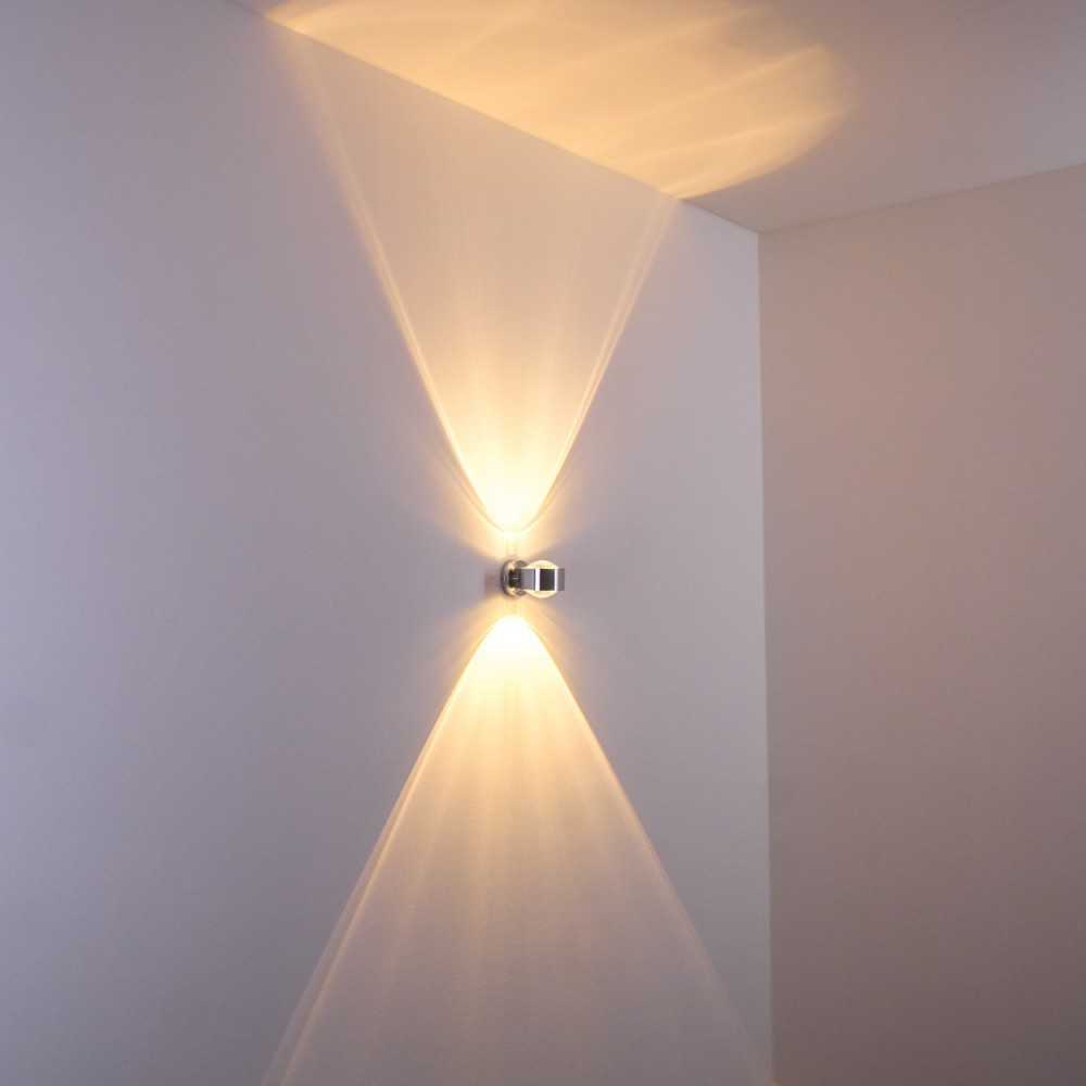 Eine moderne Wandlampe mit zwei Lichtstrahlen, die die Wand beleuchten. Die Lampe ist verchromt und an einer weißen Wand montiert.