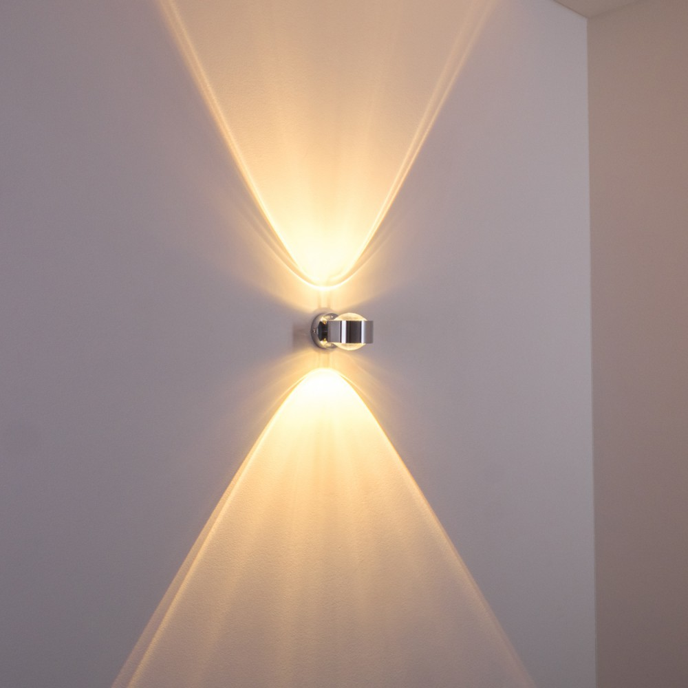 Eine moderne Wandleuchte mit Lichtstrahlen. Die Lampe ist verchromt, an einer weißen Wand montiert und wirft zwei Lichtmuster.