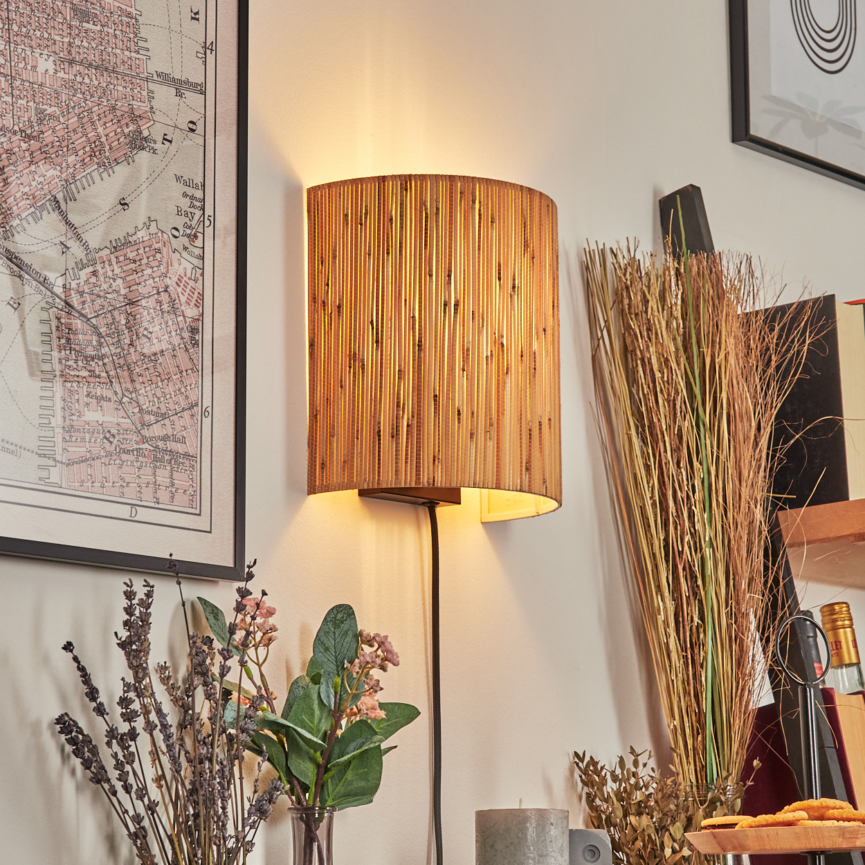 Eine Wandlampe aus Bambus leuchtet an einer weißen Wand. Eine Karte, Blumen und Dekorationen sind an der Wand und auf dem Regal.
