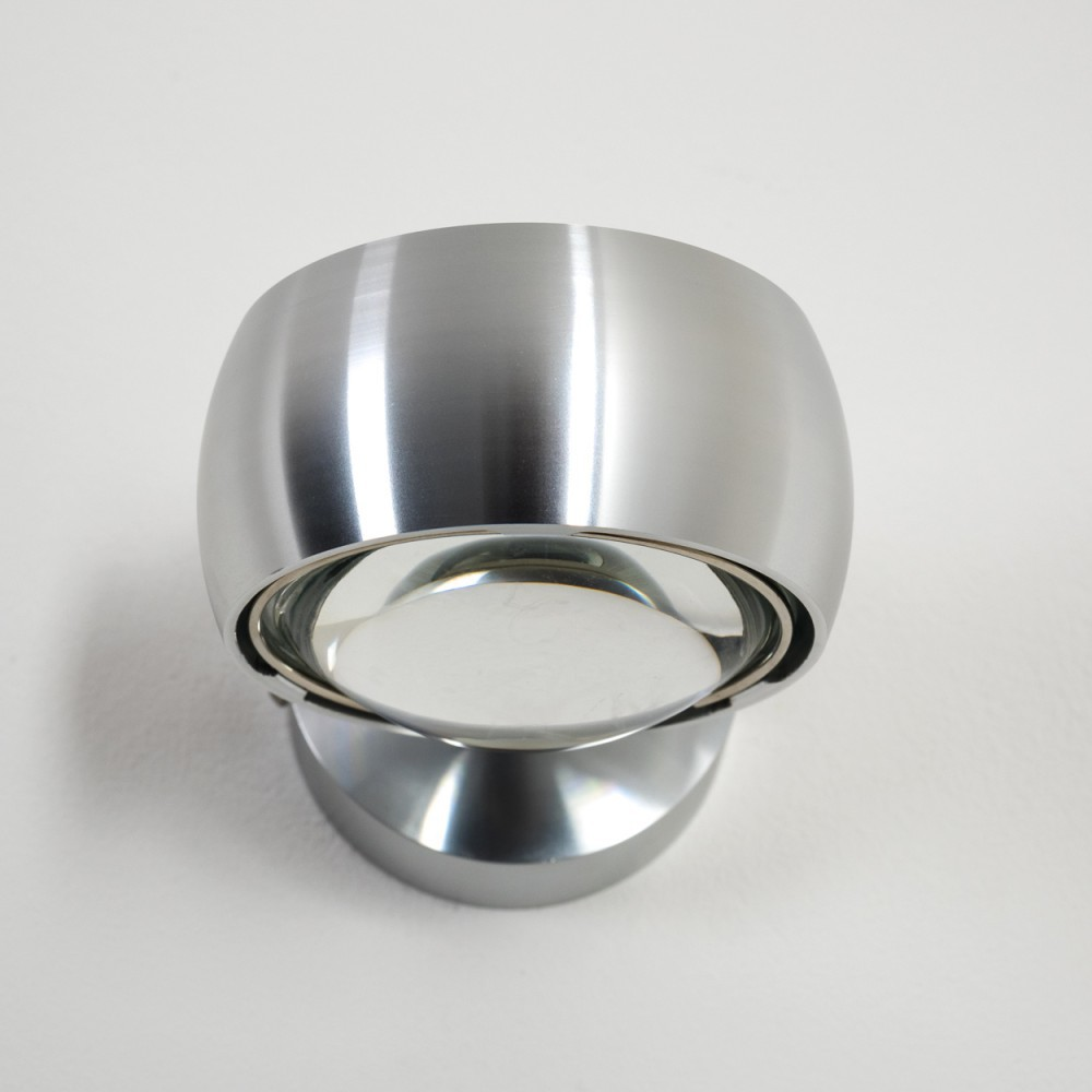 Eine metallische, moderne Wandleuchte, silberfarben, mit rundem Glaselement. Sie ist an einer weißen Wand befestigt. Die Lampe hat ein einzigartiges Design.