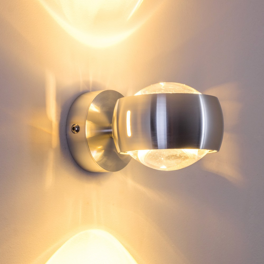 Eine moderne Wandleuchte mit silbernem Finish. Sie hat einen kugelförmigen Glasschirm und strahlt warmes Licht auf die Wand.