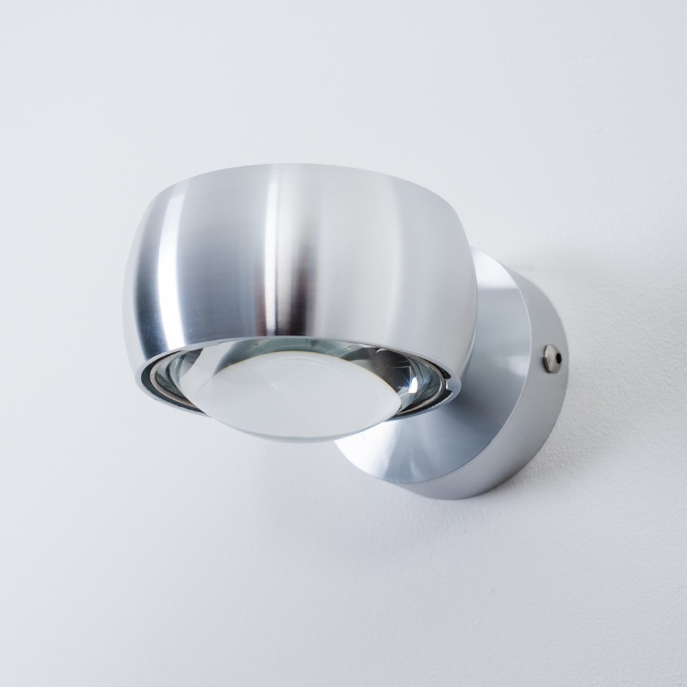 Eine moderne, metallische Wandleuchte ist an einer weißen Wand montiert. Die Lampe hat einen kugelförmigen Glasschirm und eine silberne Oberfläche.