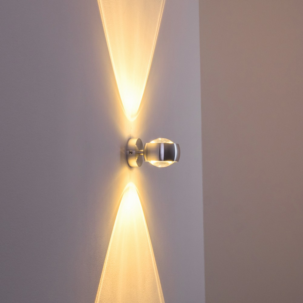 Eine moderne Wandlampe mit silbernem Sockel und Glasabdeckung. Sie strahlt Licht nach oben und unten aus und erzeugt einen Leuchteffekt.