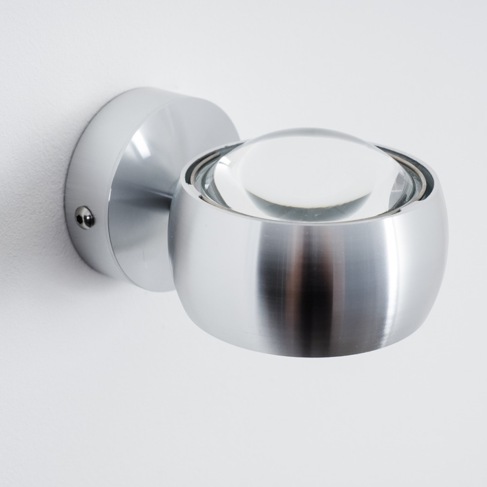 Eine moderne Wandleuchte aus Metall mit Glaslinse, an einer weißen Wand befestigt. Die Leuchte ist silberfarben und hat ein kugelförmiges Design.