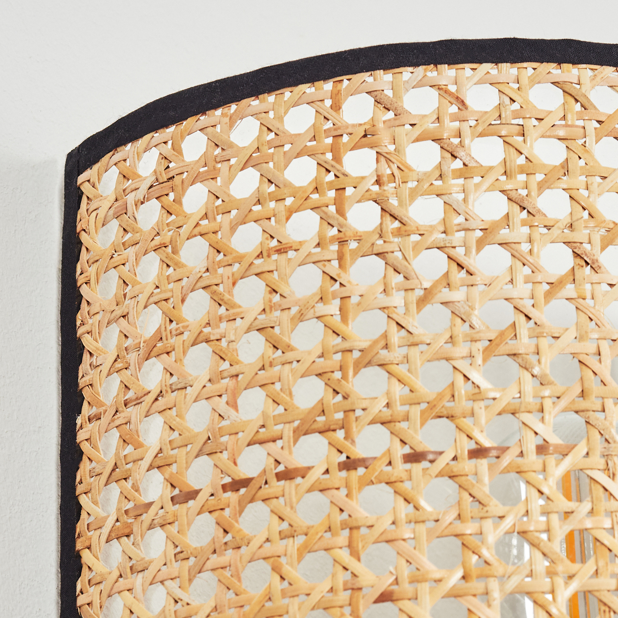 Nahaufnahme eines gewebten Rattan-Spiegels. Der Rahmen ist schwarz, der Hintergrund eine weiße Wand. Das Rattan ist hellbraun.