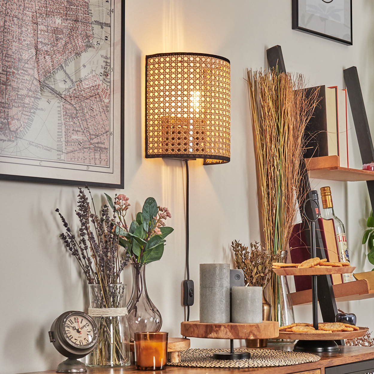 Eine beleuchtete Wandlampe aus Rattan. Eine Karte, Blumen, eine Uhr, Kerzen und Kekse stehen auf einem Holzregal.