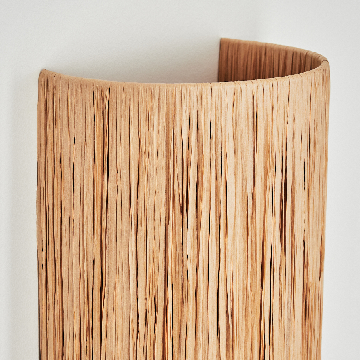 Nahaufnahme einer Wandlampe. Sie besteht aus beigem, strukturiertem Material und hat eine gebogene Form. Der Hintergrund ist eine schlichte weiße Wand.