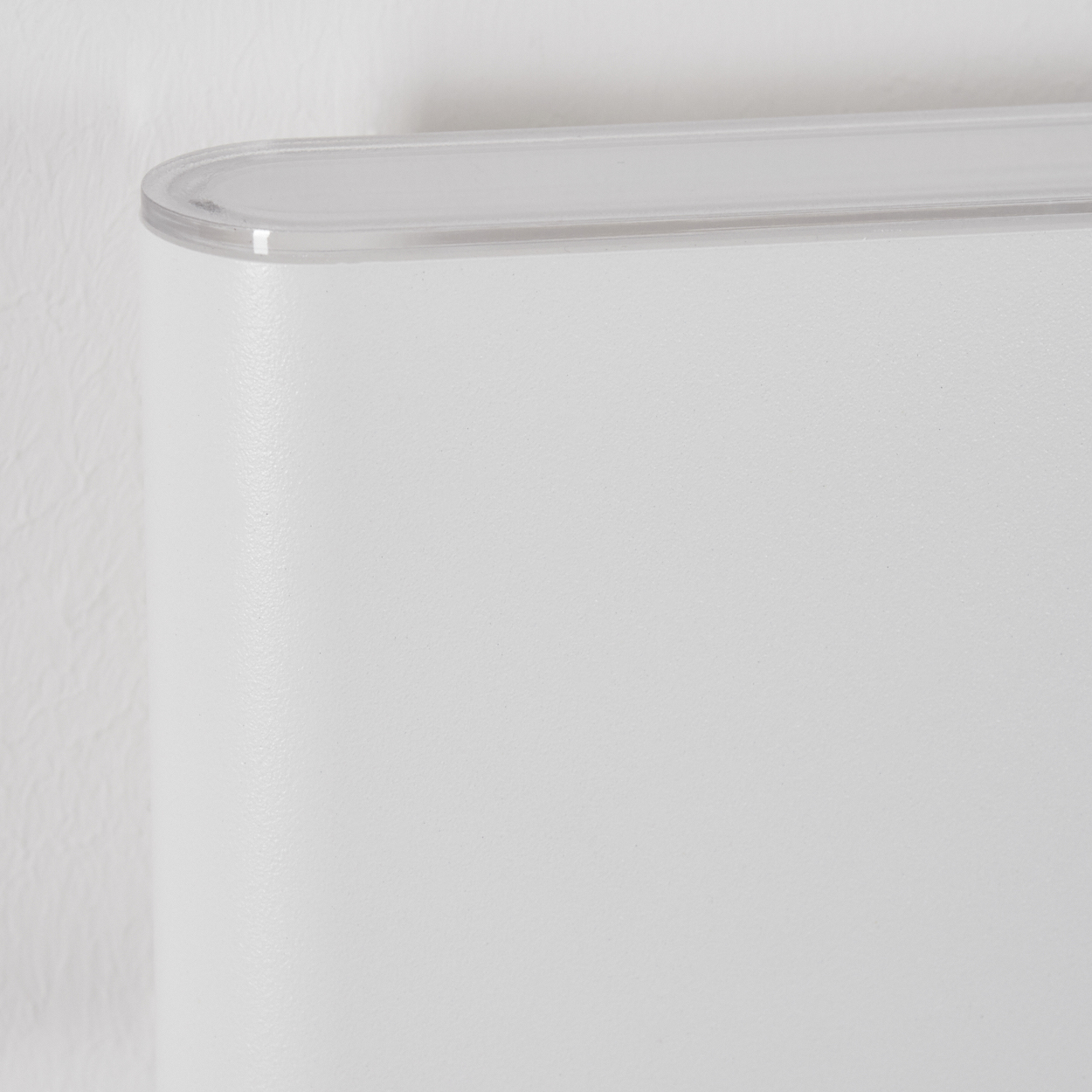 Nahaufnahme eines weißen, abgerundeten rechteckigen Objekts mit einer transparenten, gebogenen Oberseite. Es steht vor einer weißen Wand.