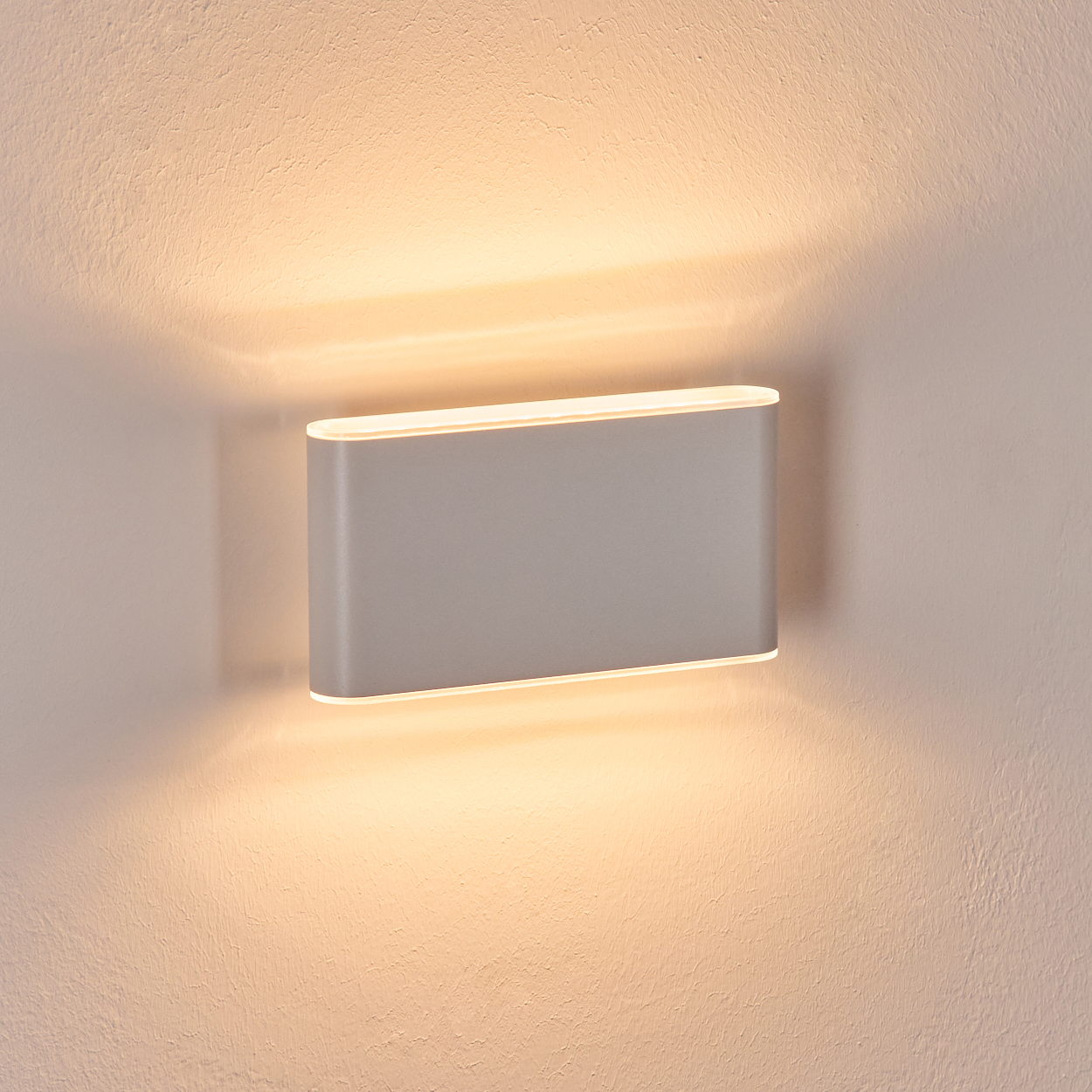 Eine moderne, rechteckige Wandleuchte ist an einer strukturierten Wand befestigt. Die Lampe strahlt ein warmes, diffuses Licht aus und wirft Schatten.