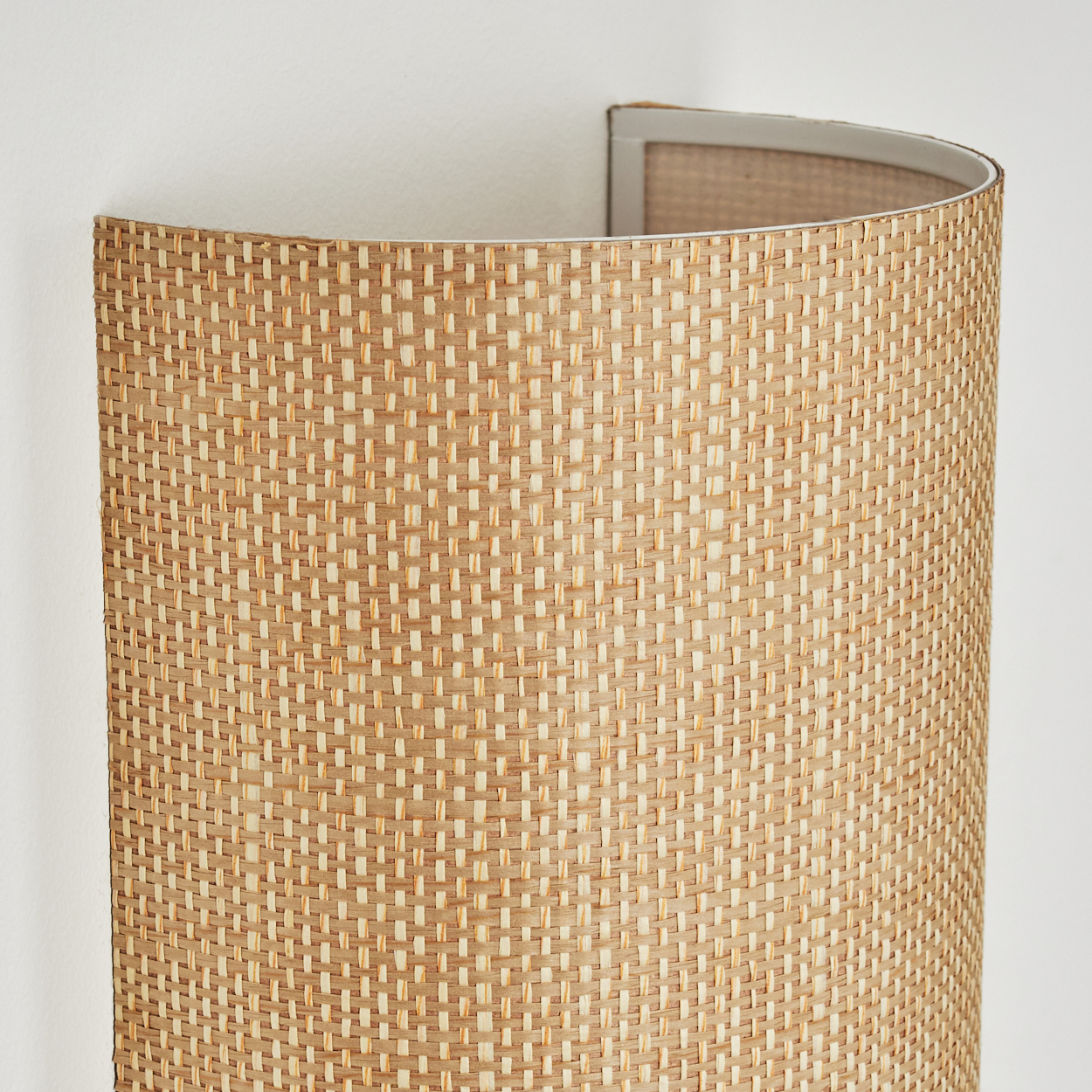 Eine beige gewebte Wandleuchte. Die Lampe hat eine gebogene Form und ist an einer weißen Wand montiert. Das gewebte Material ist sichtbar.