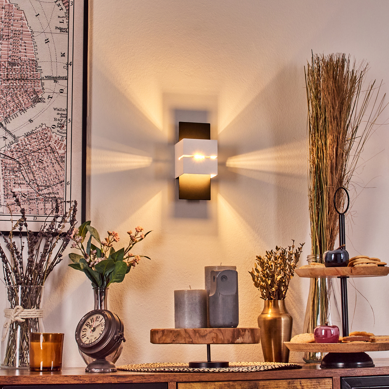 Eine Wandlampe strahlt Licht aus. Auf einem Holztisch stehen Kerzen, Blumen und eine Karte. Eine Glasvase und eine Uhr sind ebenfalls zu sehen.