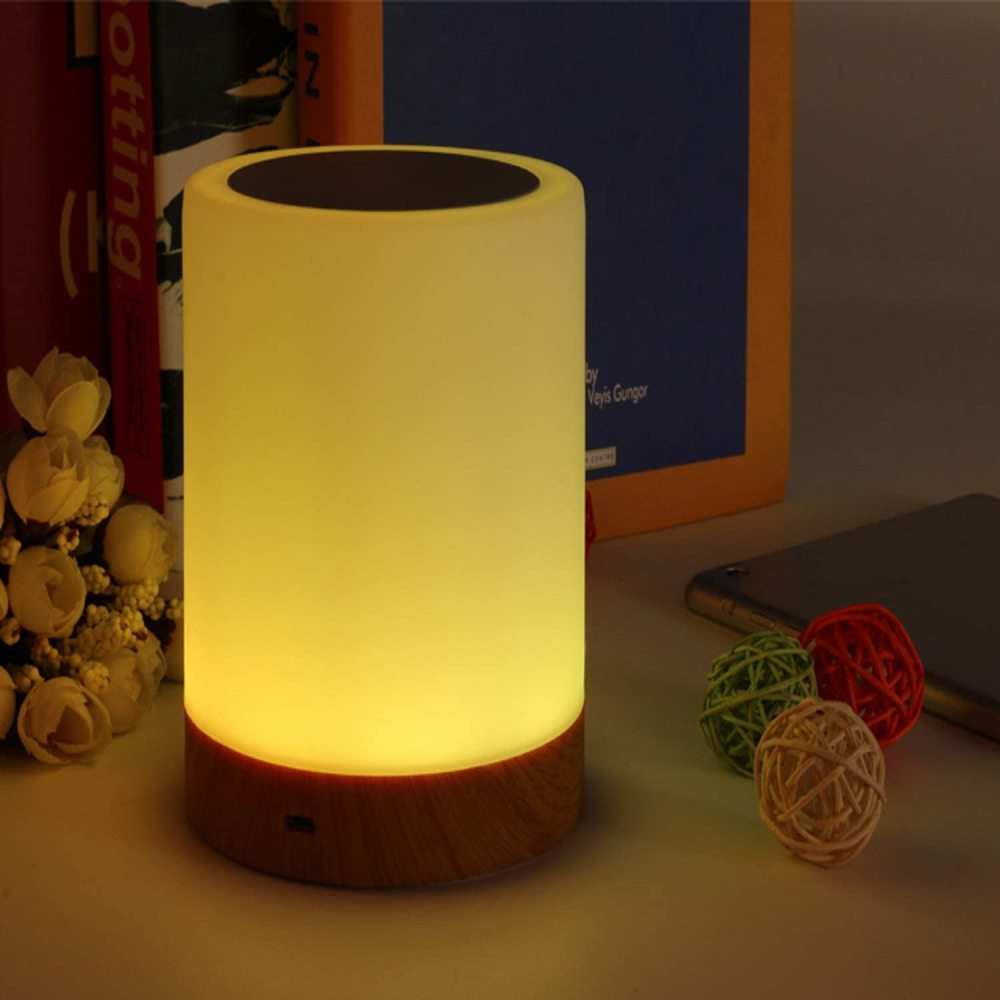 Eine leuchtende zylindrische Lampe mit Holzsockel. Daneben liegen dekorative Kugeln und ein Smartphone. Bücher sind im Hintergrund.