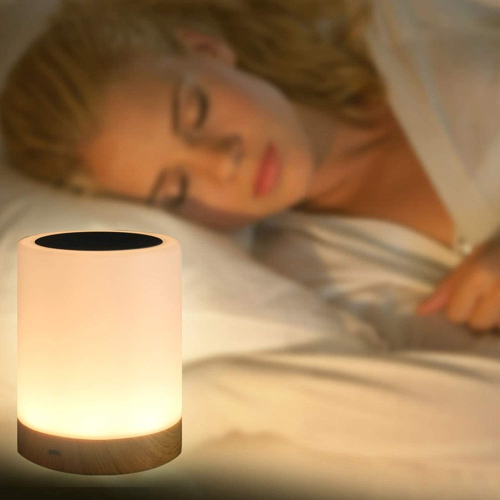 Eine leuchtende Nachttischlampe mit einer schlafenden Frau im Hintergrund. Die Lampe ist zylindrisch, mit Holzsockel und sanftem Schein.
